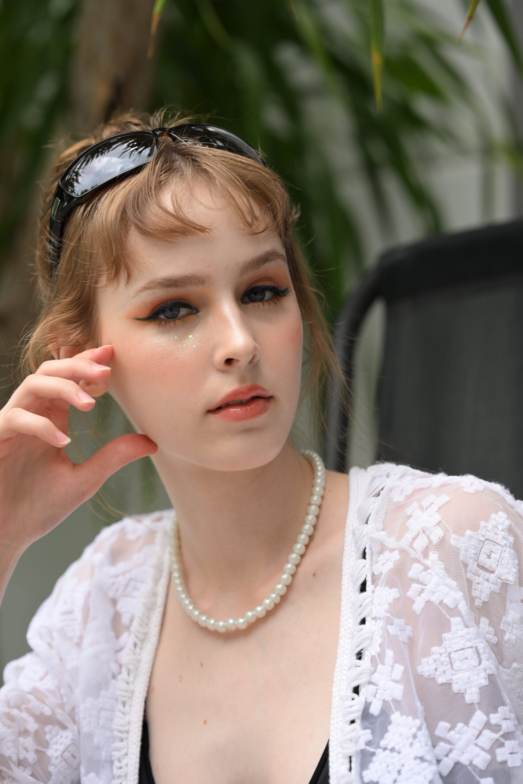 A woman with a pearl necklace và sunglasses smiles, showcasing a final dewy skin makeup look with a bold eyeliner and shimmering glitter.