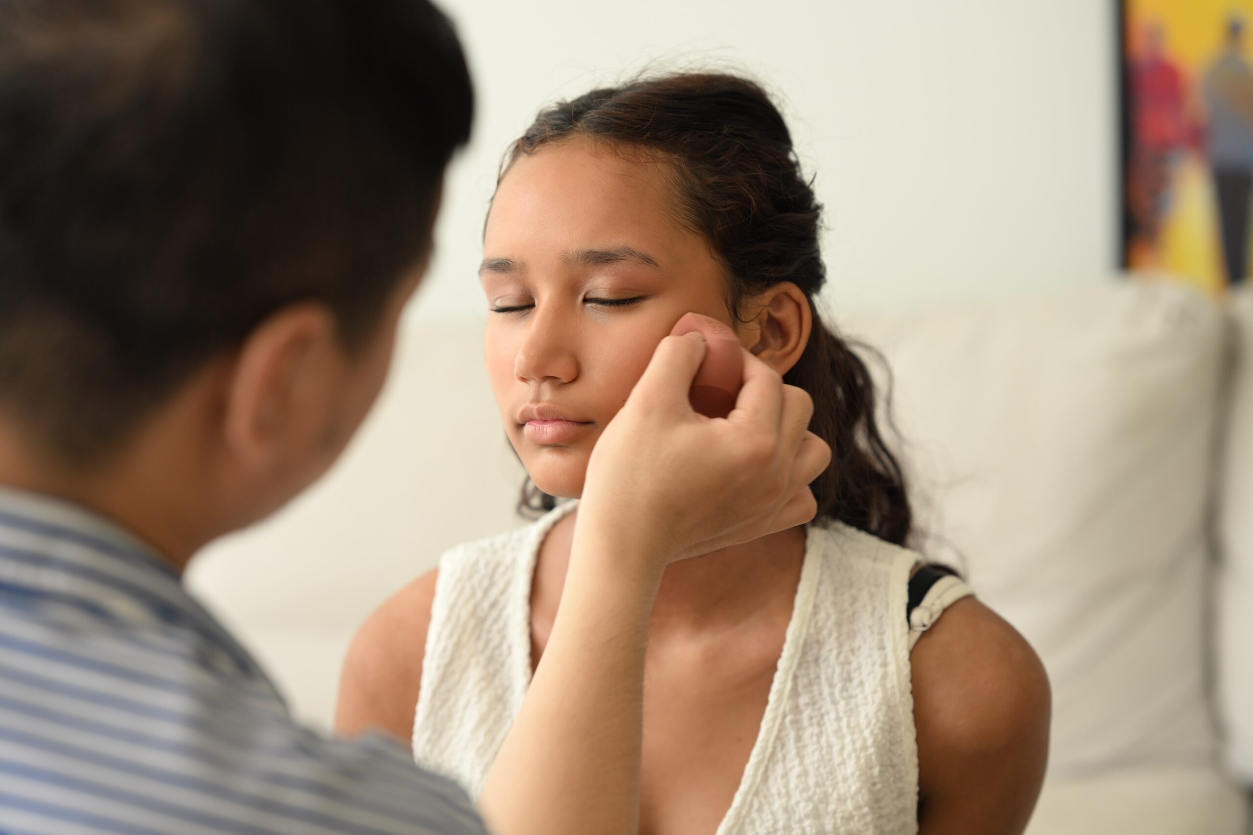 A makeup artist applies product with a sponge to a woman's face, demonstrating a step in a dewy skin makeup tutorial for a radiant finish.