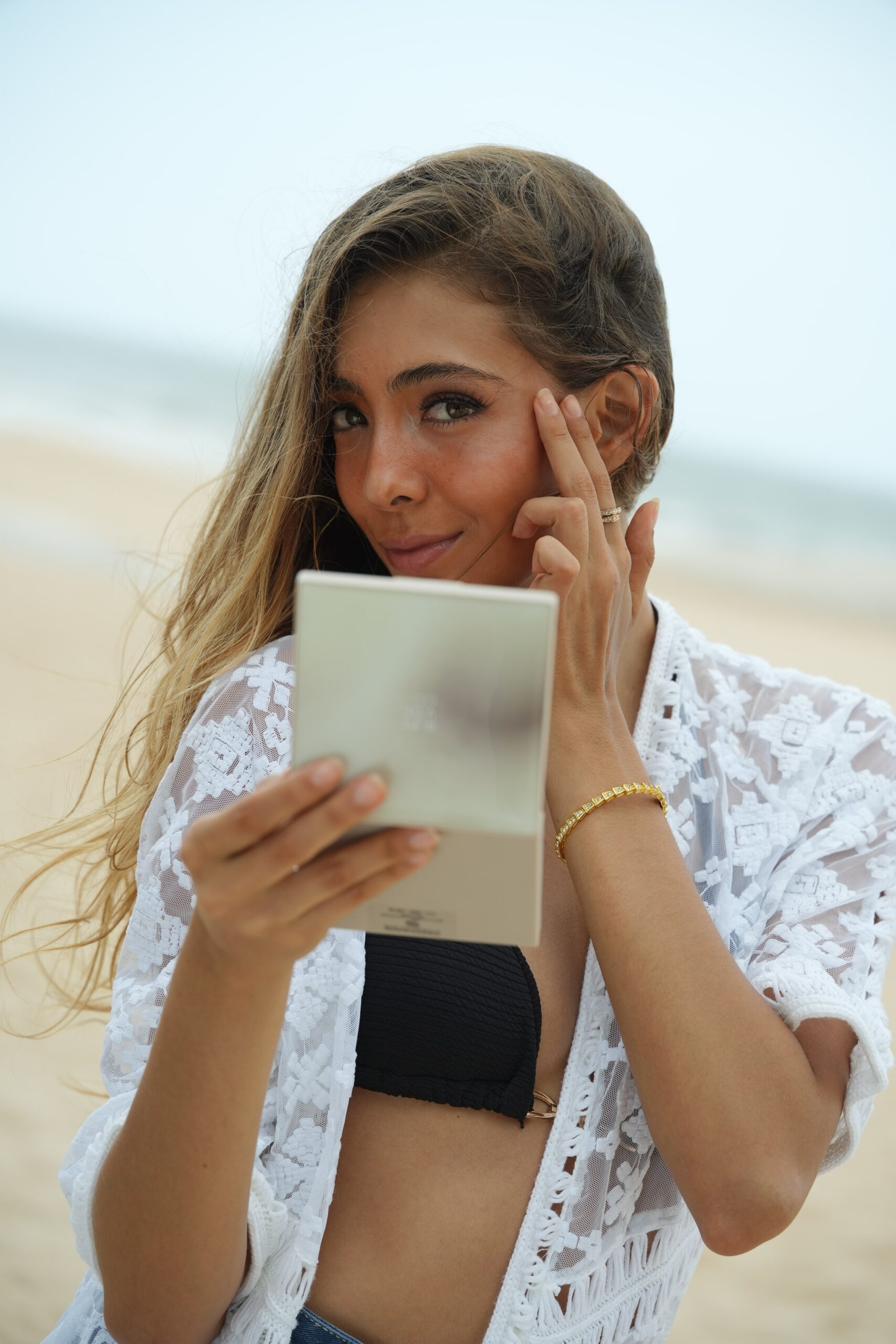 A woman looks into a compact mirror while applying makeup on a beach, showcasing a routine for a radiant, dewy skin glow.