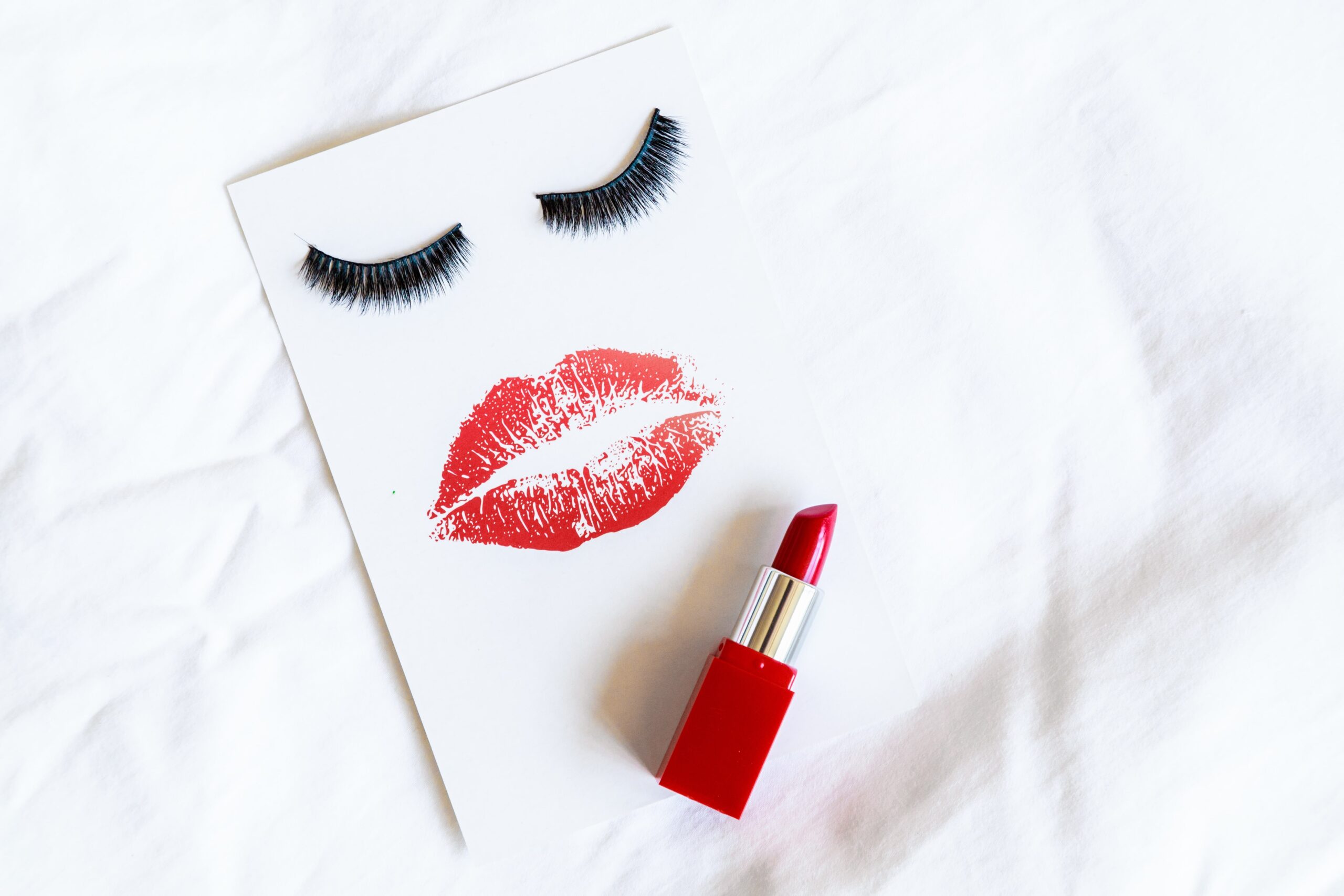 A flat lay of a red lipstick with a lipstick print and false eyelashes on a white background, representing the elements of a Taylor Swift-inspired makeup look.