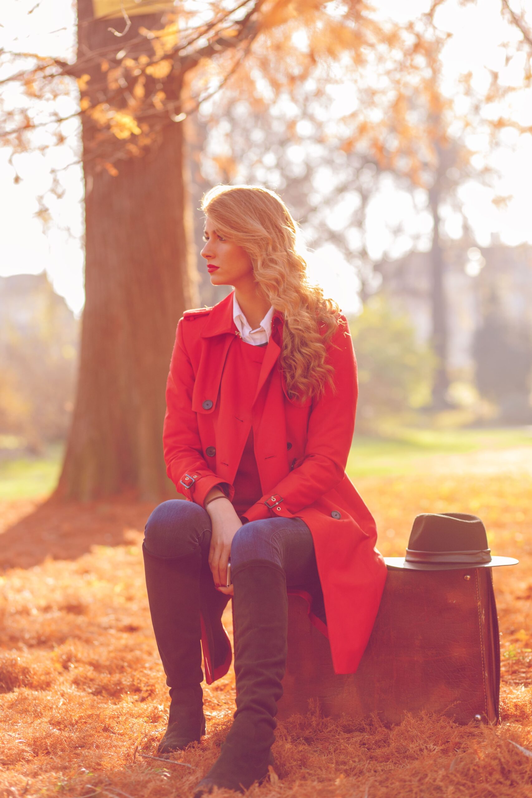 A woman with a bold red lip wears a red coat and sits on a suitcase in an autumn park, showcasing a classic look inspired by Taylor Swift.