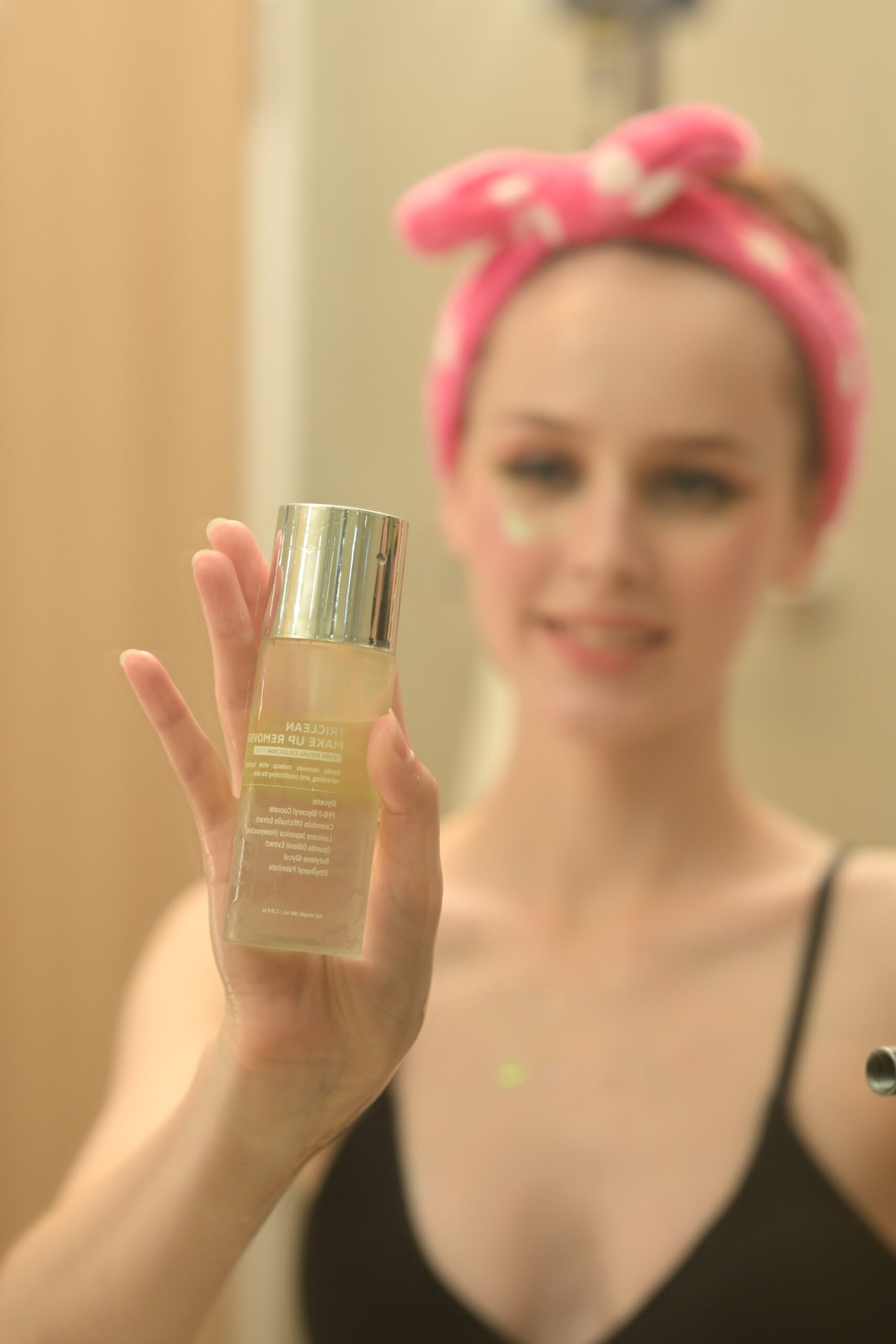 A woman with a pink headband smiles while holding a bottle of makeup remover, representing a key step in a cleansing routine for glowing skin.