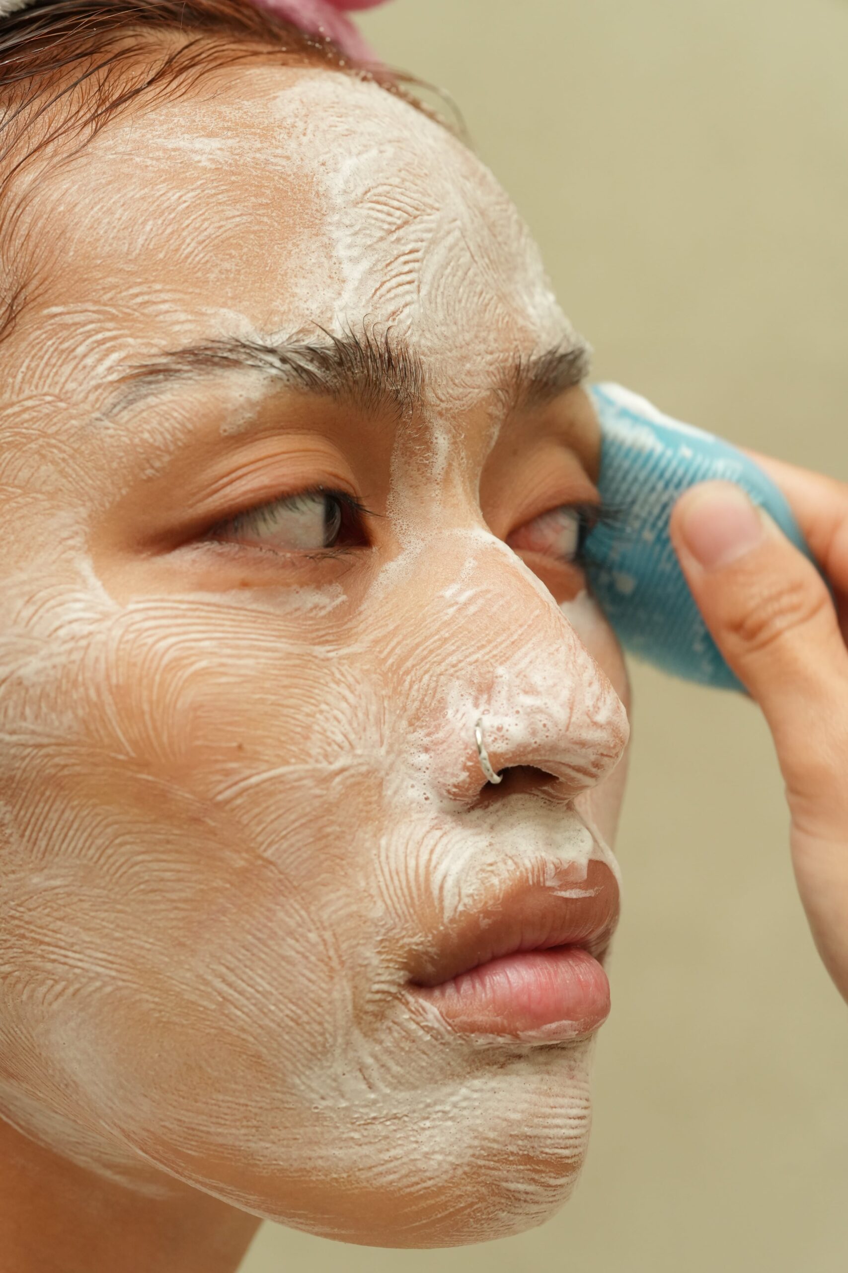 A person uses a blue facial cleansing brush to massage foamy cleanser onto their face, a step-by-step guide on how to use a cleanser right for glowing skin.