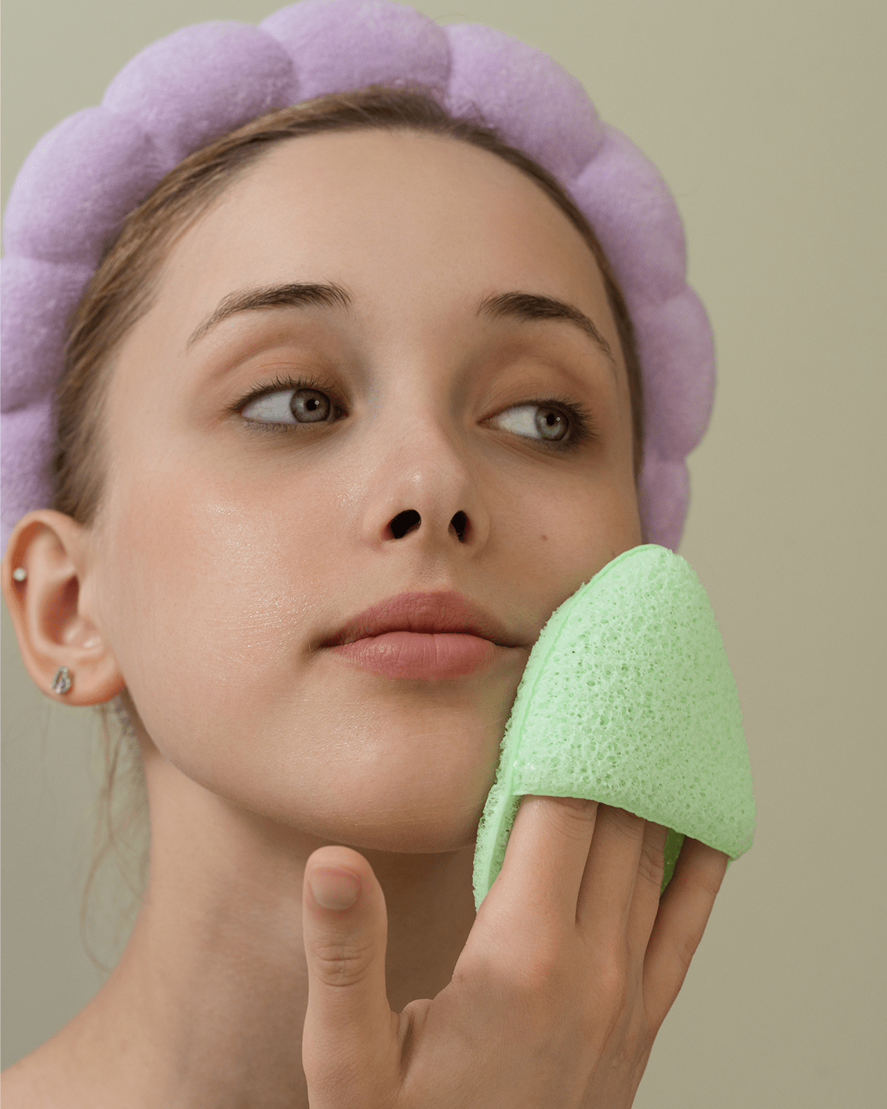 A woman with a lilac headband uses a green sponge-like pad to gently cleanse her face, promoting a perfect cleansing routine.