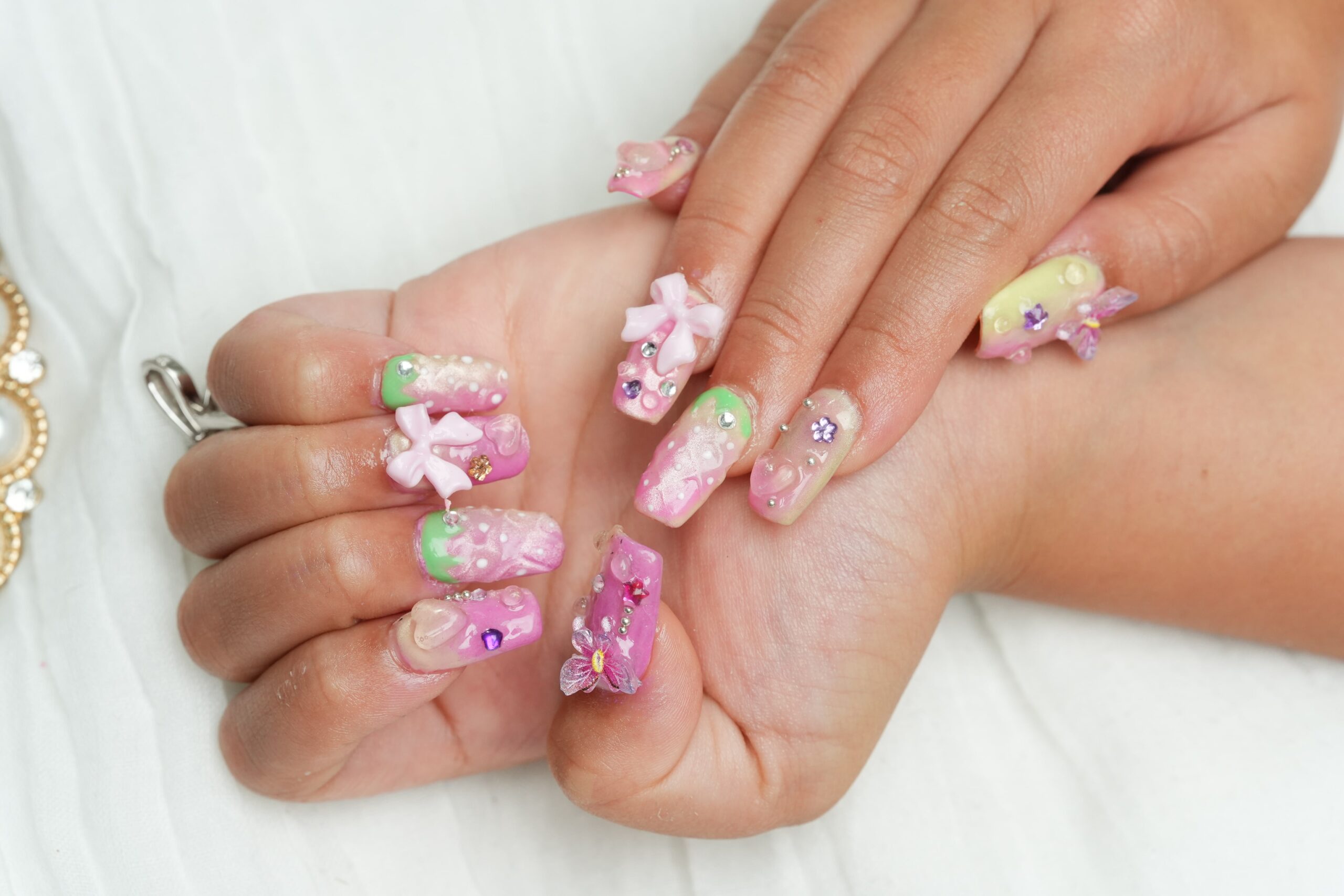 A detailed shot of a person's hands with intricate, decorated nails. The nails feature a gradient of neon pink and green, adorned with 3D bows, rhinestones, and gems, showcasing a bold and elaborate nail art style.