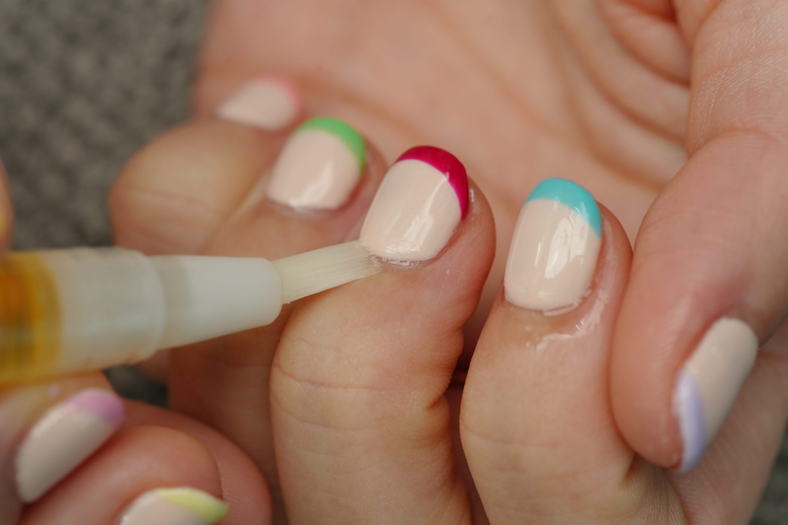 A person applying cuticle oil to a nail with a convenient pen applicator. The surrounding nails feature a modern French manicure style with bright neon tips in fuchsia, mint green, and turquoise, highlighting the importance of nail care for a flawless manicure.