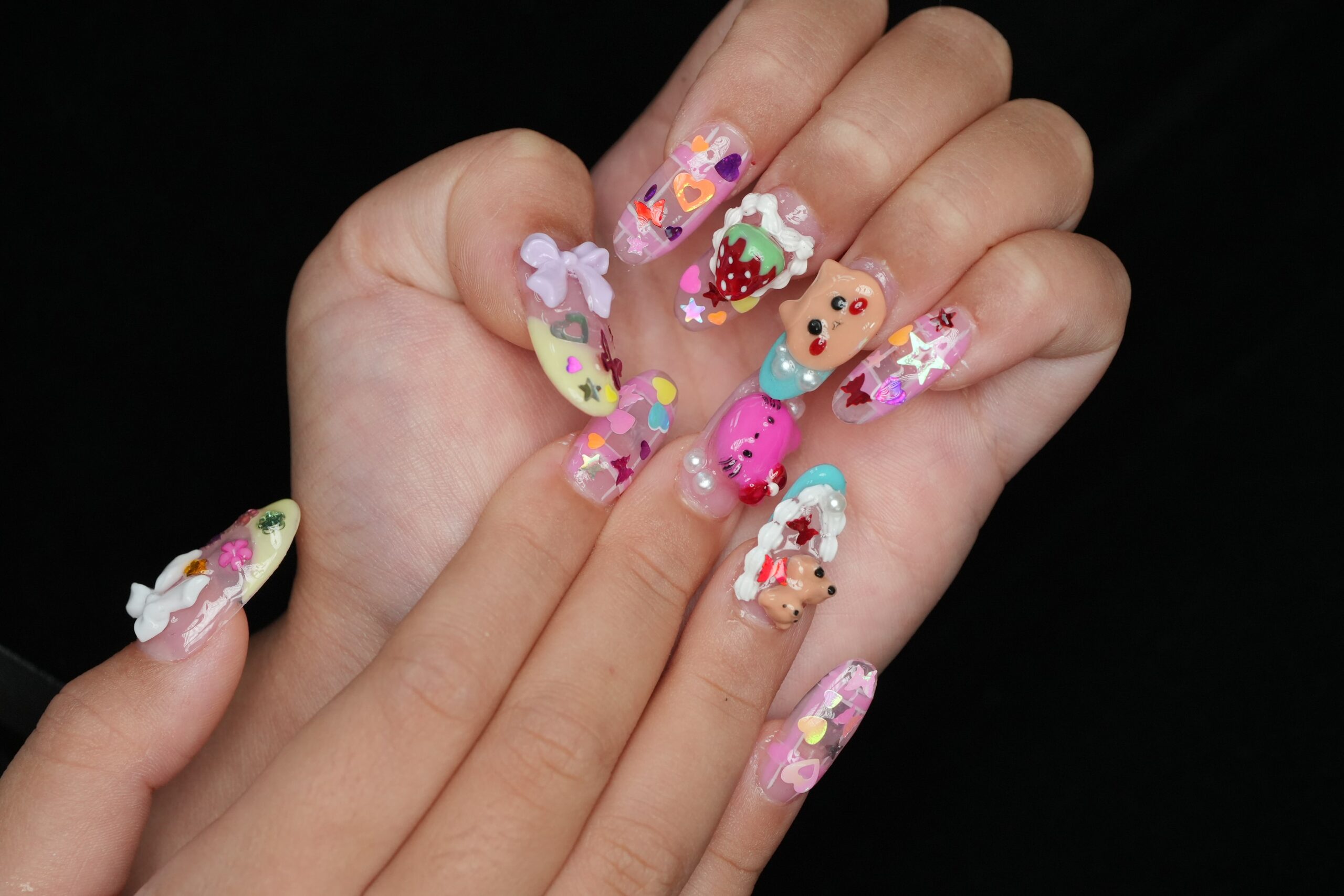 A hand with a collection of highly decorated false nails featuring playful 3D designs. The nails have a clear base with various summer-themed charms, including hearts, a teddy bear, strawberries, and bows, all in vibrant neon and pastel colors.