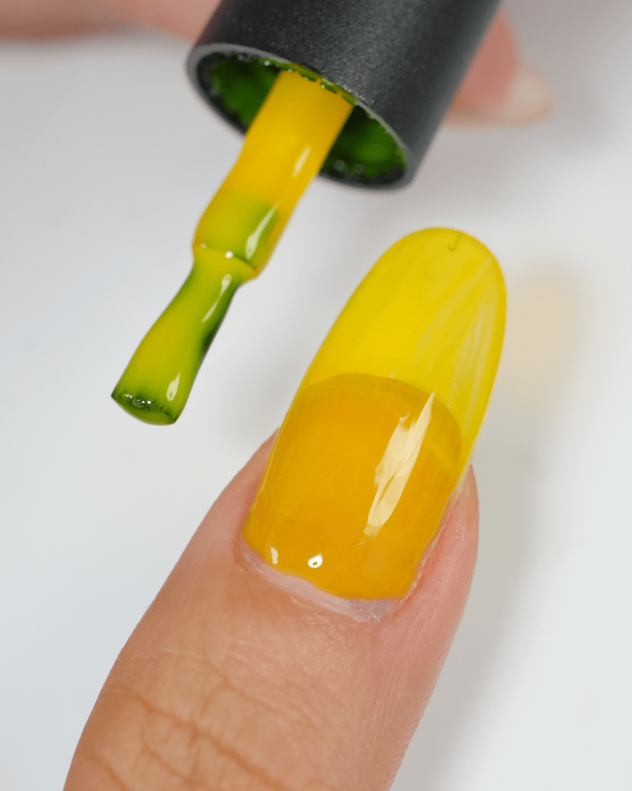 A close-up shot of a person applying a bright yellow gel polish on their nail with a brush. The image shows the first coat of the neon yellow color being applied over a clear base, highlighting the bold pigment.