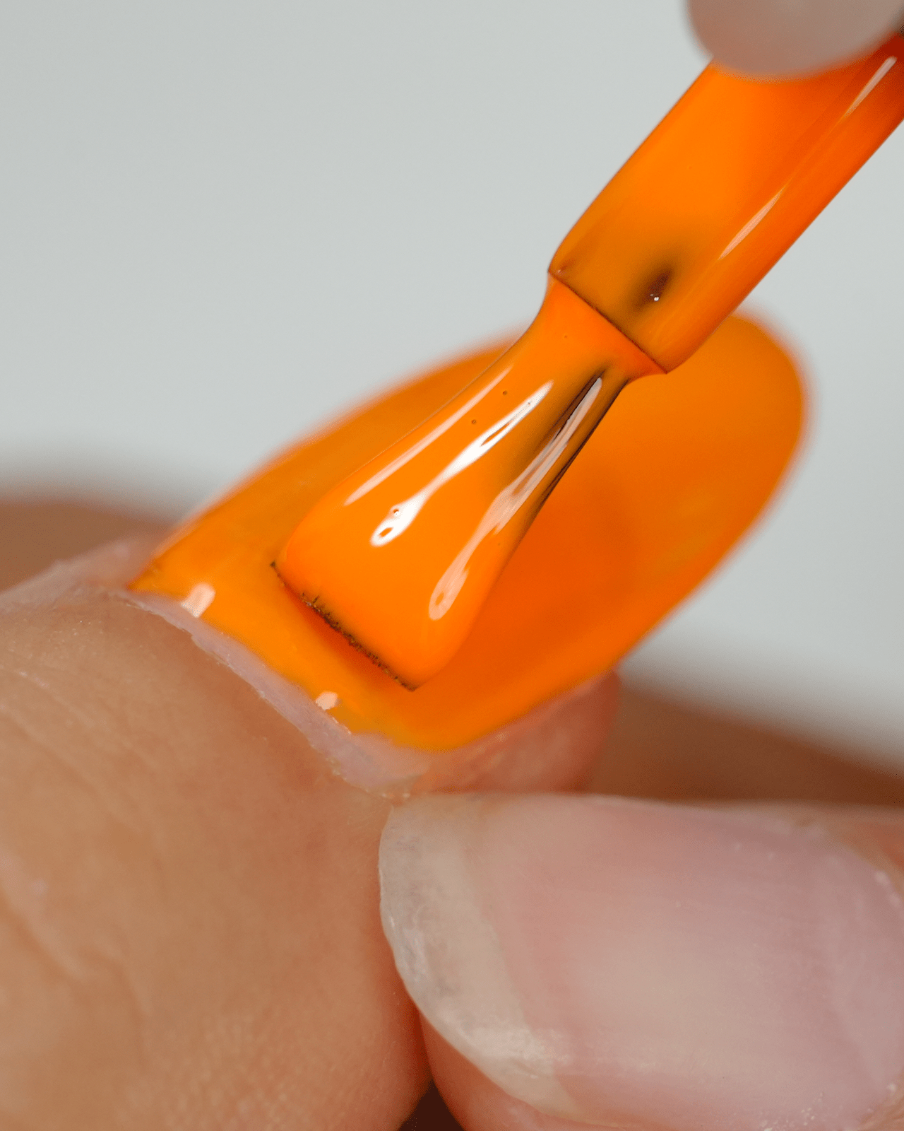 A close-up of a person painting their nail with a bright orange gel polish, showcasing the smooth application and high-shine finish of the neon color.