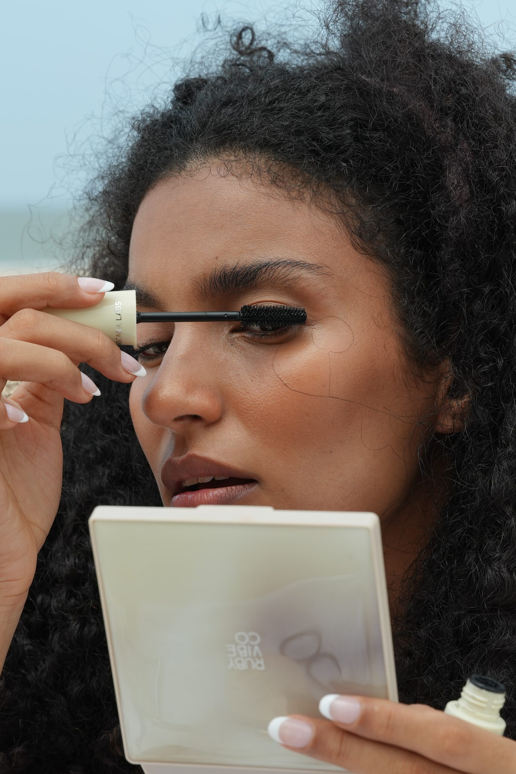 A woman with curly hair applies mascara while holding a compact mirror, demonstrating a makeup step that pairs well with a nude lip