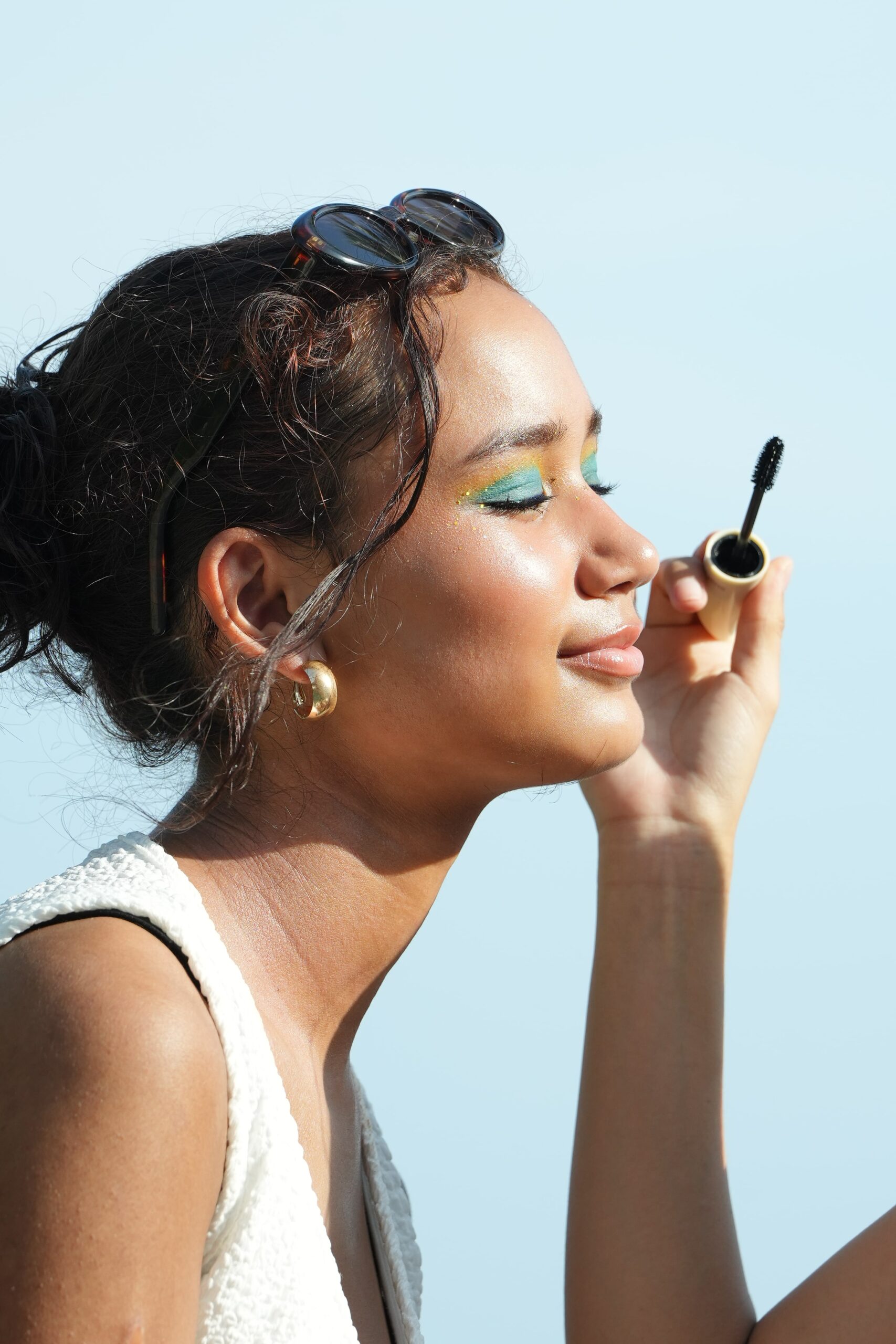 A woman with sunglasses applies mascara, with a bold eyeshadow look, showing a makeup step that pairs well with a nude lipstick for any occasion.