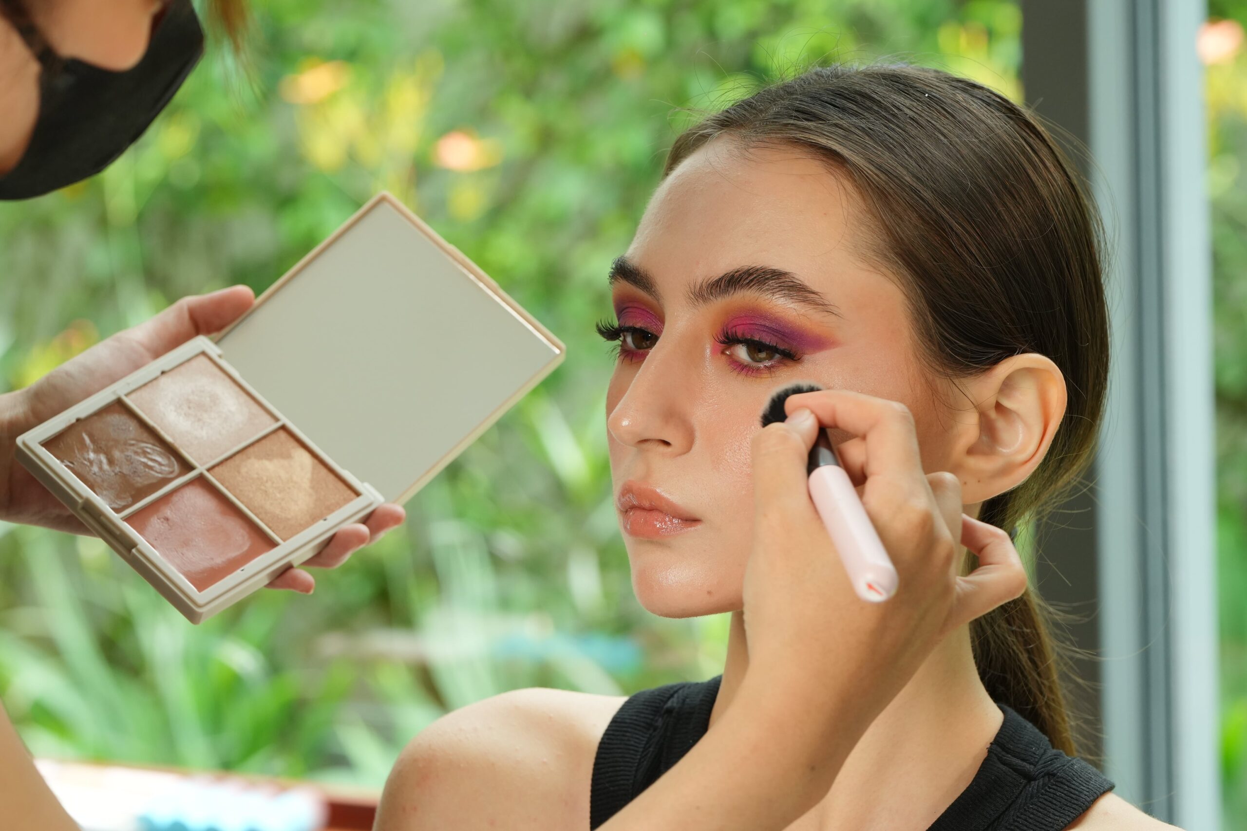 A person applies a vibrant eyeshadow to a woman's eyelid, showing a step in a makeup tutorial focused on achieving a smooth and fresh finish.