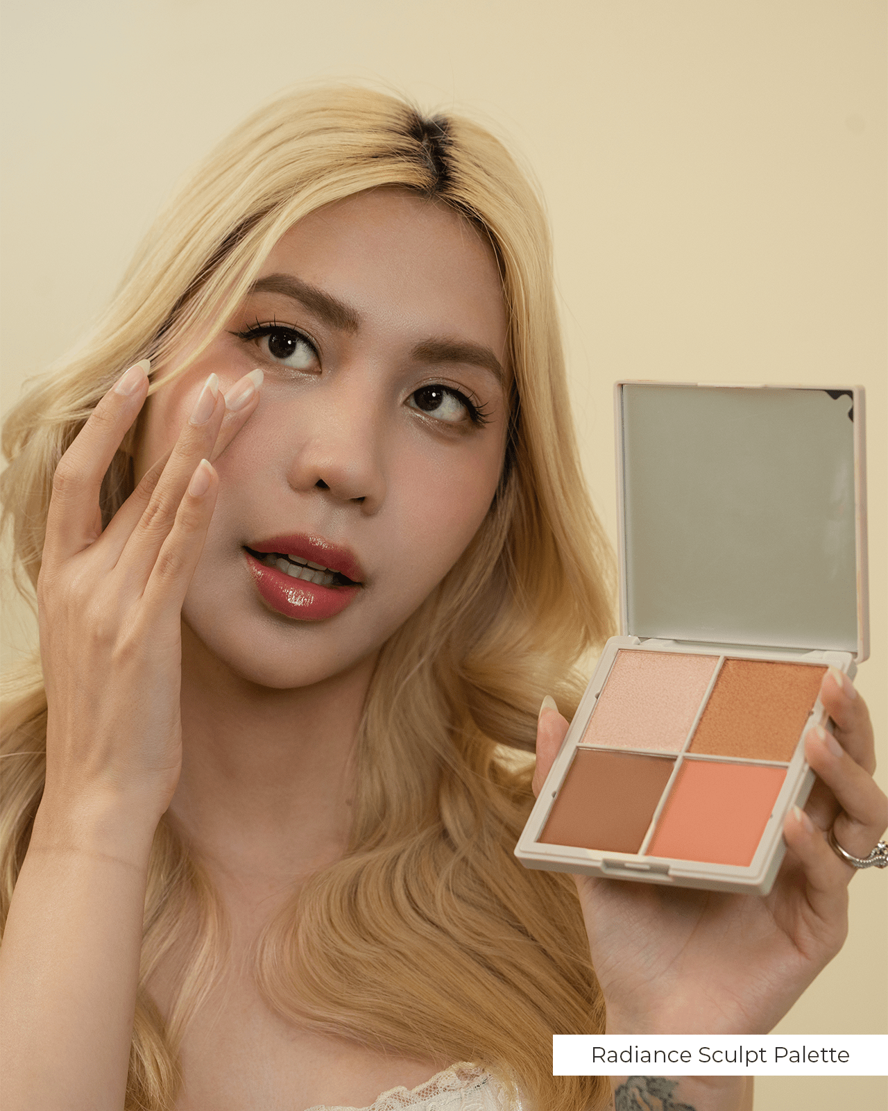 A blonde woman holding a makeup palette named "Radiance Sculpt Palette." She is applying makeup with her finger, showcasing a full-face look. This photo emphasizes the broader makeup routine that complements a bold lip color.