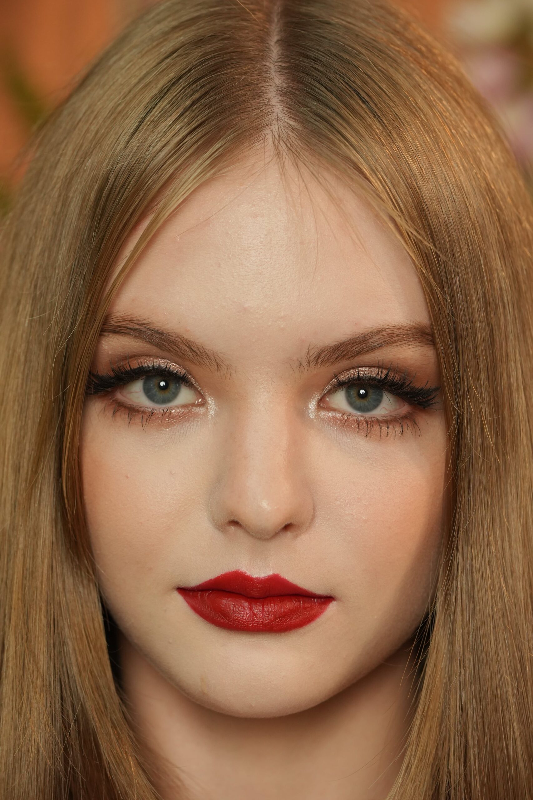  A close-up, straight-on portrait of a woman with long blonde hair and blue eyes, wearing a striking red lipstick and winged eyeliner. The image focuses on her confident expression and the flawless application of the red lip.