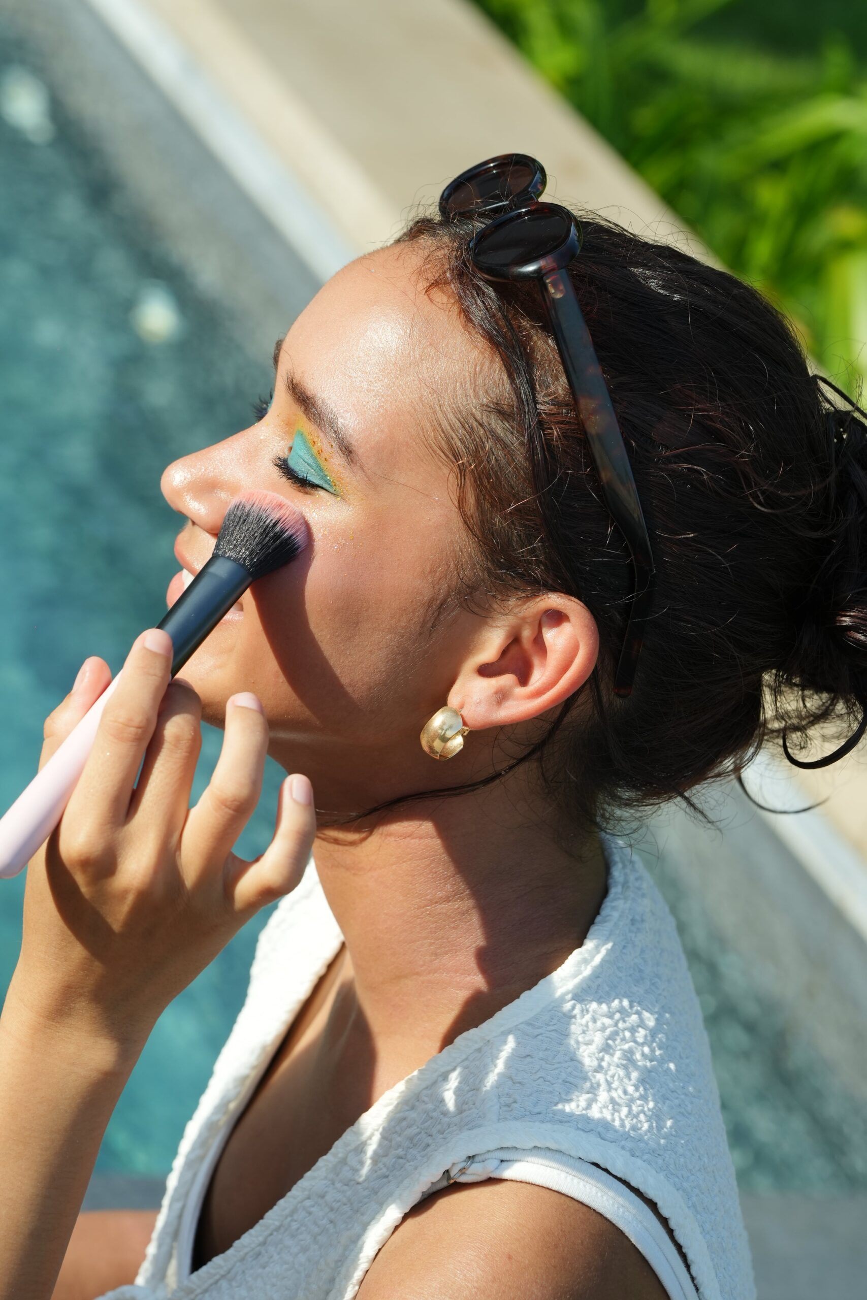 A woman applies blush to her cheek with a brush, showcasing a colorful eye makeup look, as part of a makeup tutorial for beginners.