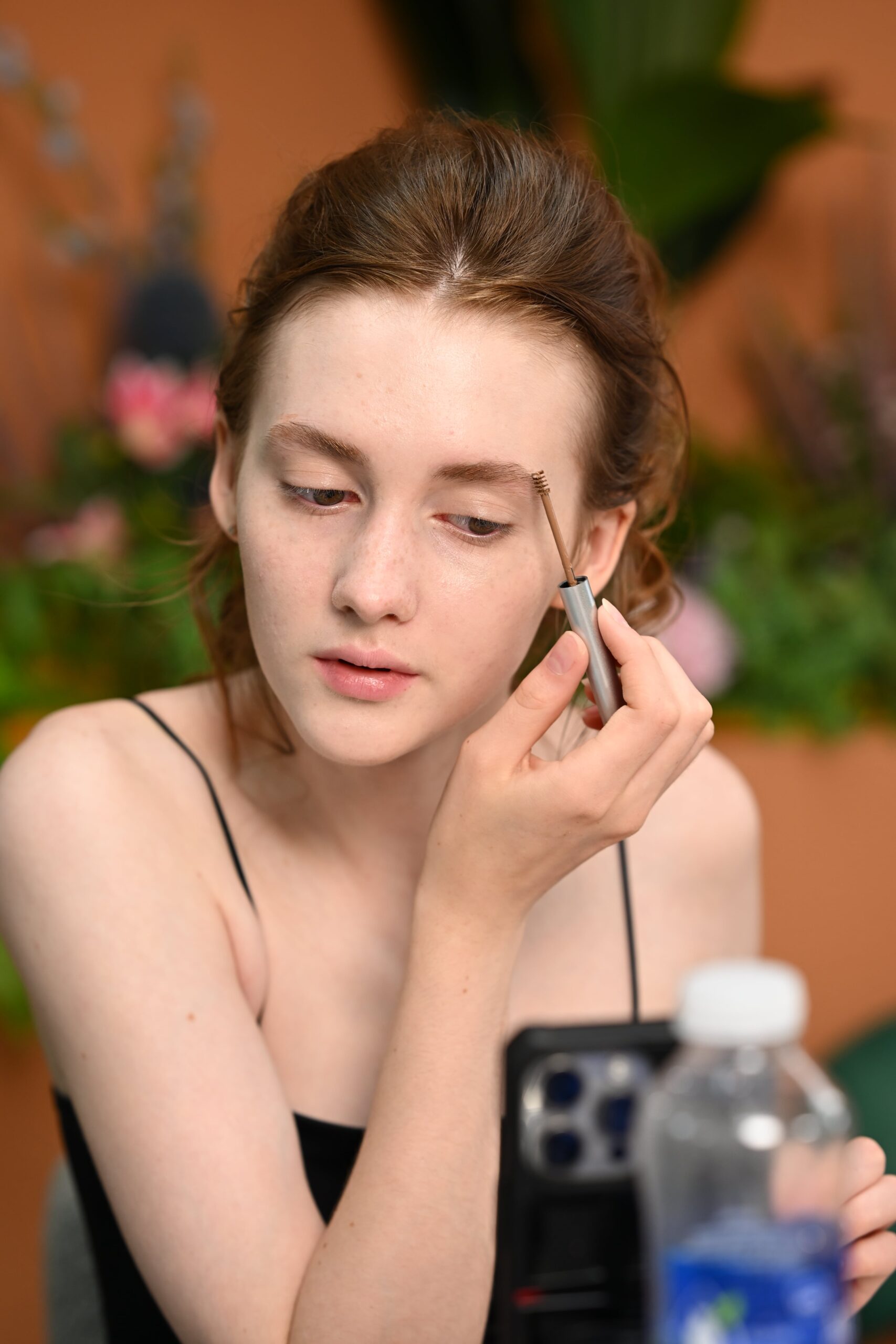 A woman applies brow gel to her eyebrows while looking at a phone, following a step-by-step makeup tutorial for beginners.