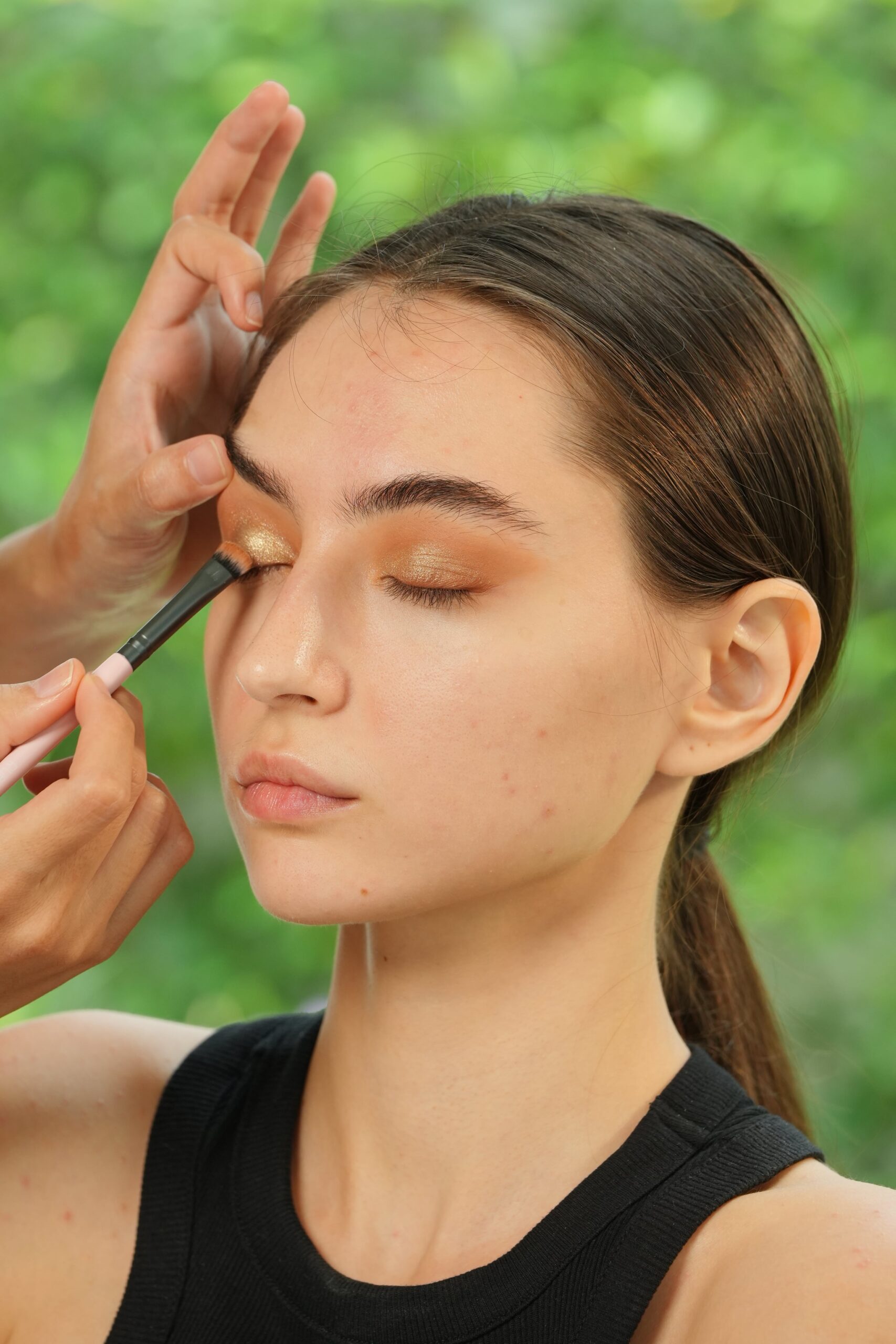 A makeup artist applies a shimmering gold eyeshadow to a woman's eyelid with a brush, showing a key step in a makeup tutorial for beginners.