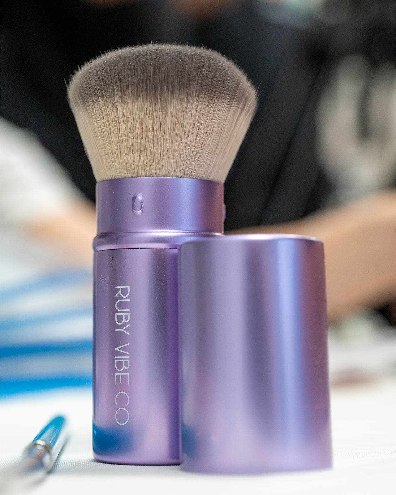 A close-up of a purple, retractable kabuki brush from the brand 'Ruby Vibe Co,' a compact tool for a teen makeup starter kit.