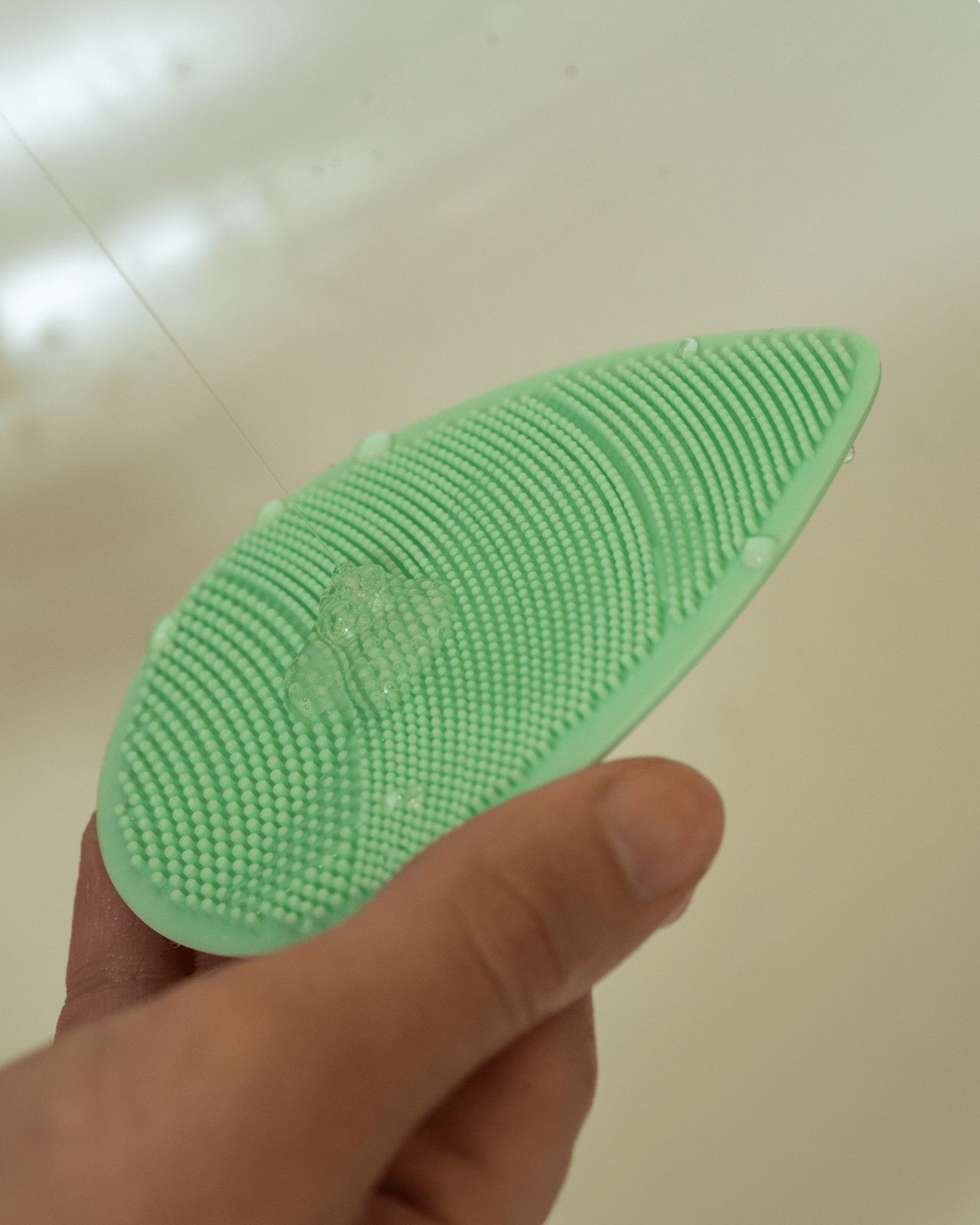 A hand holding a green silicone facial cleansing brush with water droplets on it, a must-have tool for a deep and effective cleanse.