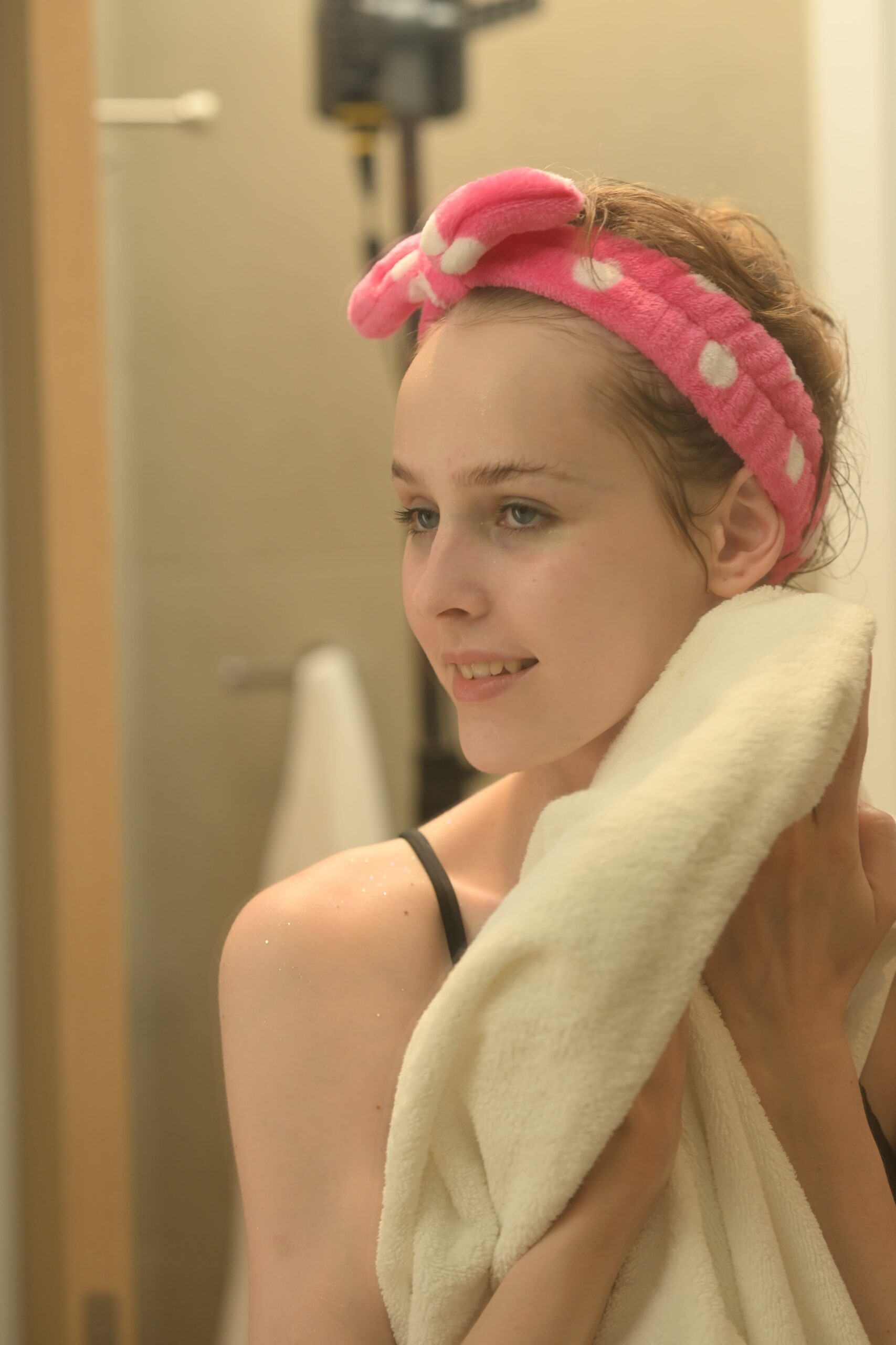 A person wearing a pink polka-dotted headband with a bow, holding a white towel and smiling while looking in the mirror, representing a fresh and clean skincare routine. The background shows a bathroom setting with a towel rack and a hairdryer. Perfect for the guide "The Right Cleanser for Your Skin: A Guide for Skin Types.