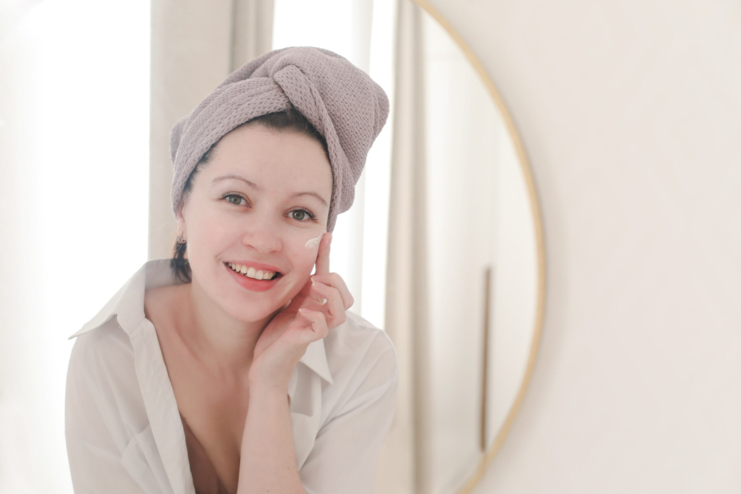 A smiling woman with clean, healthy skin applies a moisturizer after cleansing, showing the importance of a complete skincare routine for a glowing complexion.