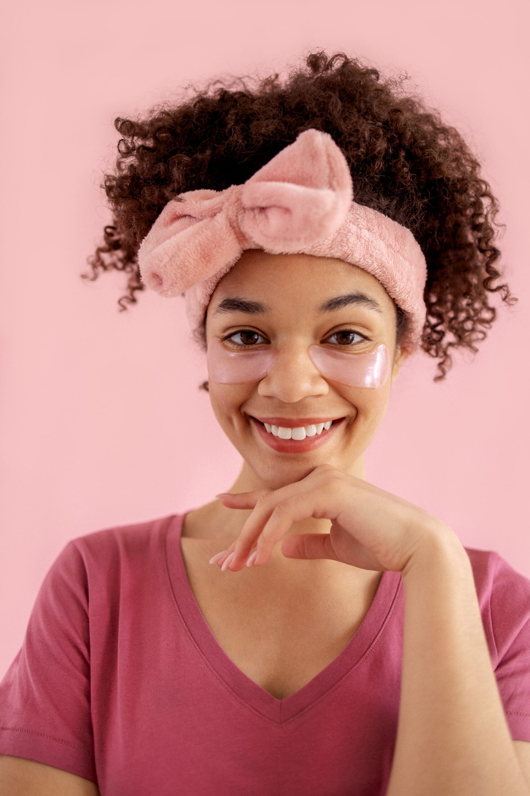 A smiling woman with a pink bow headband and eye masks, showcasing a fun and effective accessory for a skincare routine.