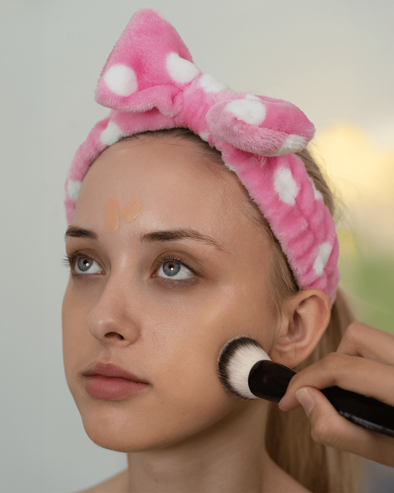 A woman with a pink polka-dot headband applying foundation with a brush, showing the usefulness of a headband during makeup application.