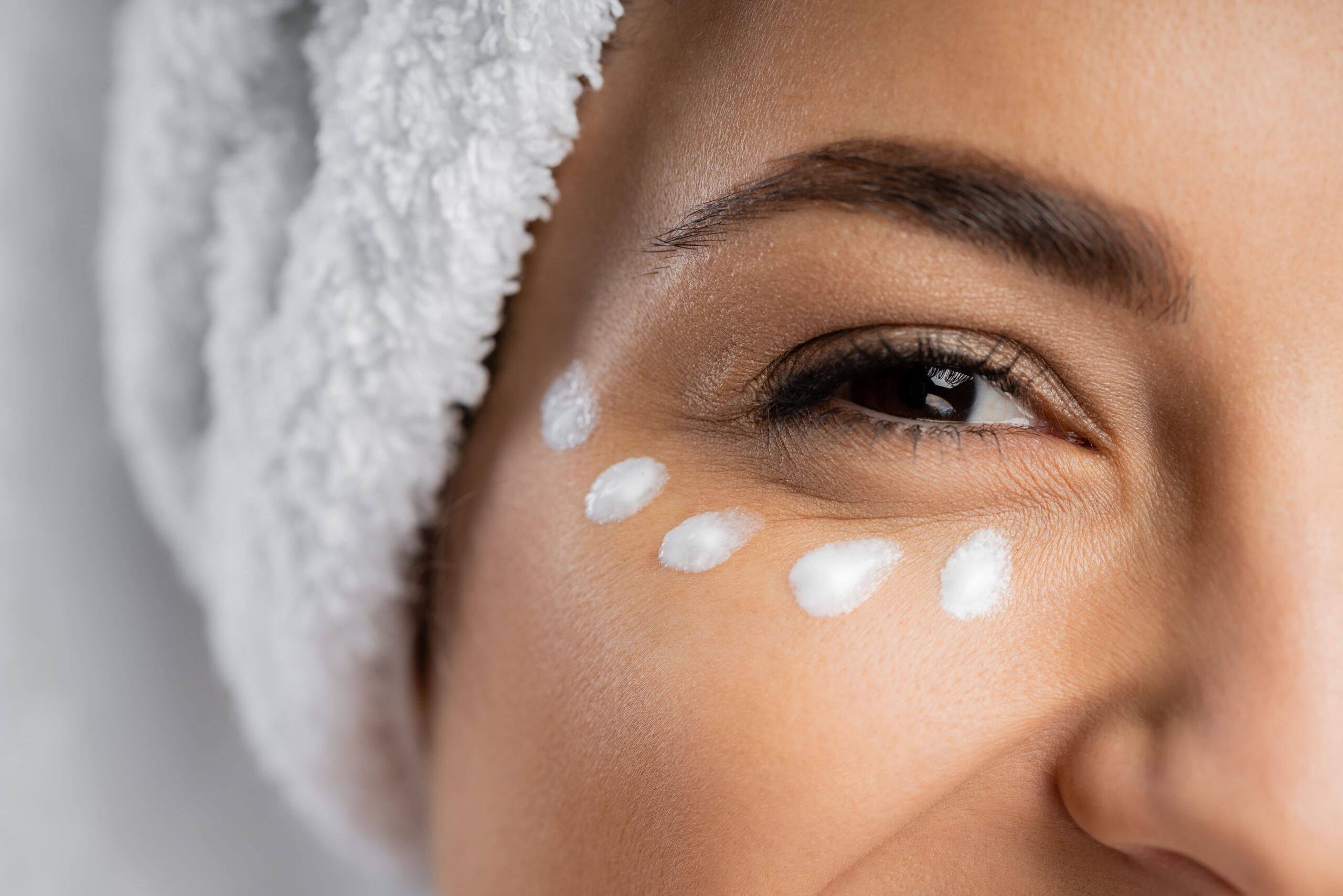 A close-up of a woman's eye area with dots of white cream applied under her eye, showing the correct application method for a retinol eye cream.