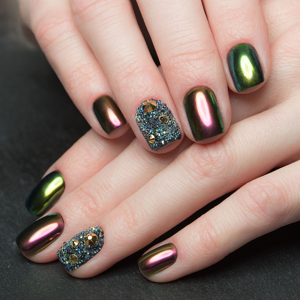A sophisticated manicure with multi-colored metallic nails that transition from green to copper, along with two nails accented with textured gemstones.