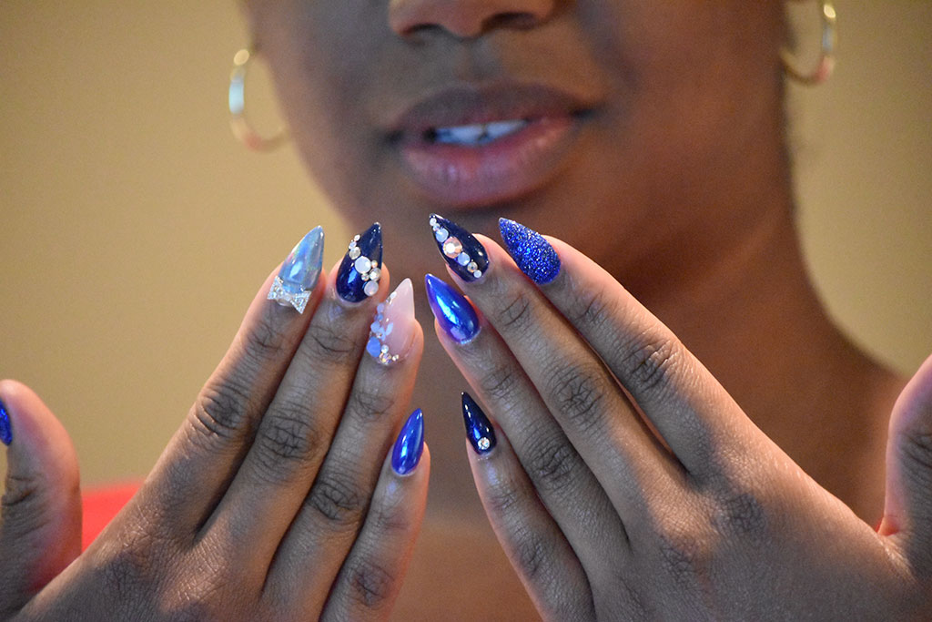 A woman shows off an elaborate blue stiletto nail look that features a combination of chrome nails, glitter, and gemstones.