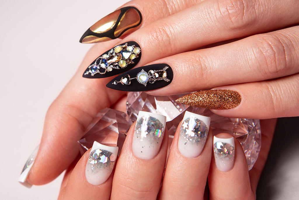 Two contrasting manicures are displayed together: one with long black pointed nails studded with gemstones, and the other with short white nails designed with glitter.