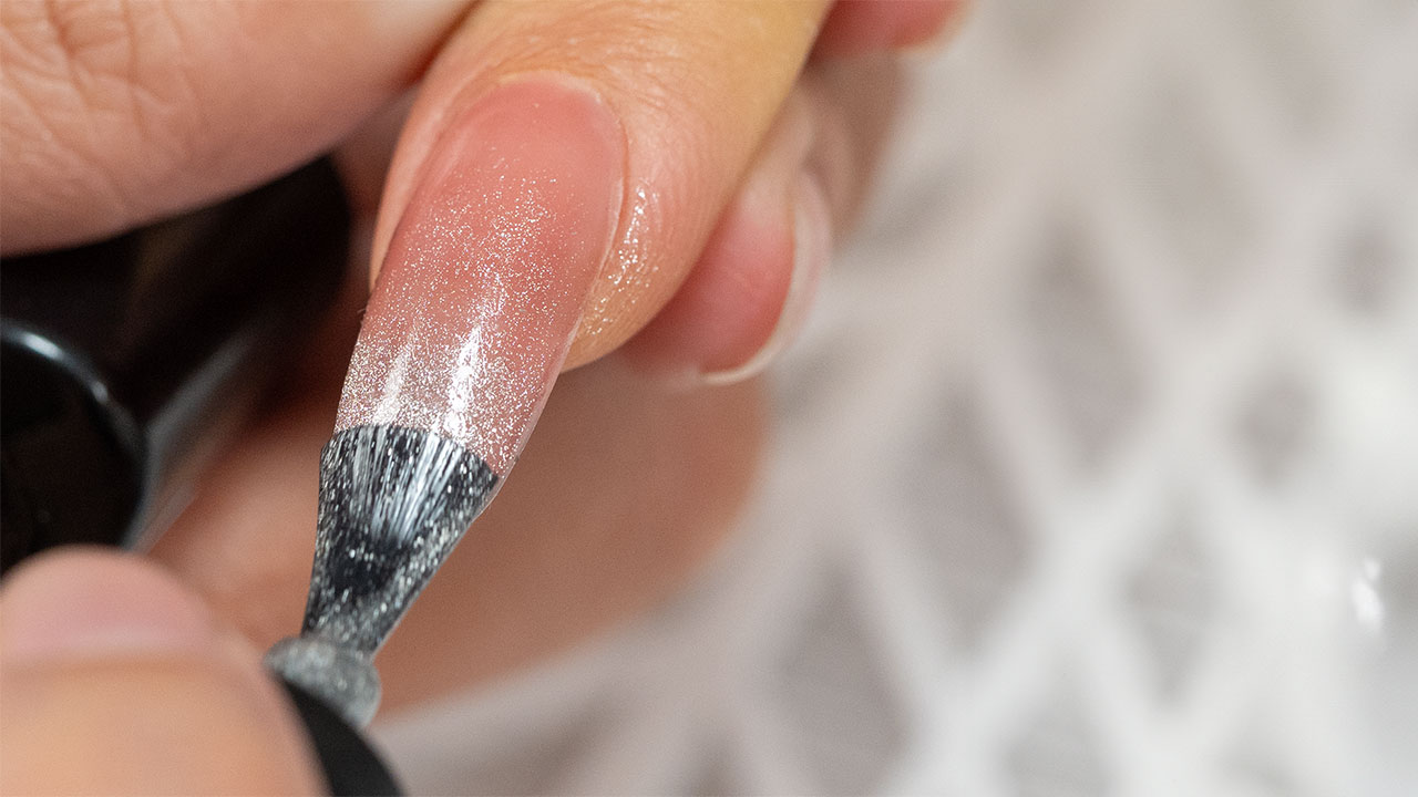 An extreme close-up of a glitter top coat being applied with a brush to a long, elegant fingernail.