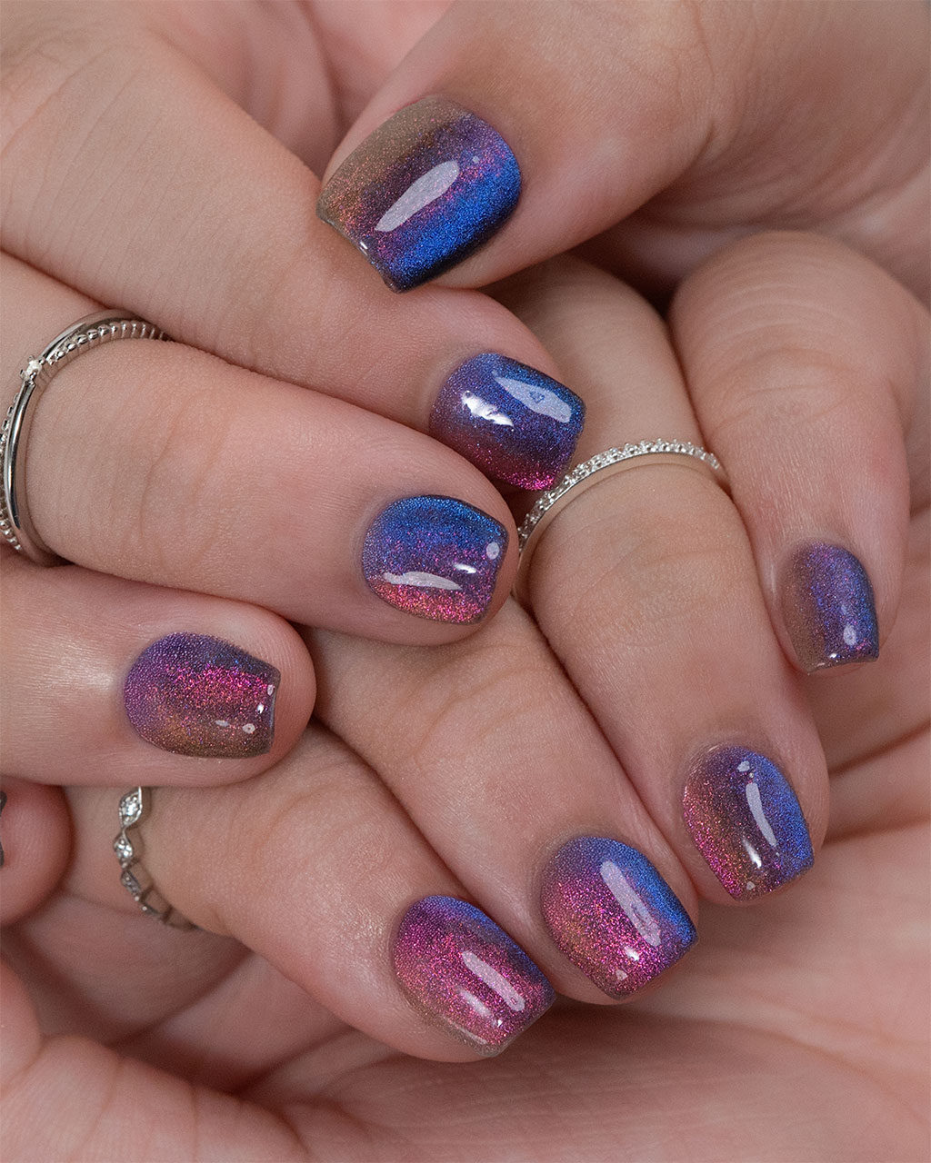 A close-up of a hand with mesmerizing multi-chrome ombre nails that shift in color from blue to purple and pink, accented with silver rings.