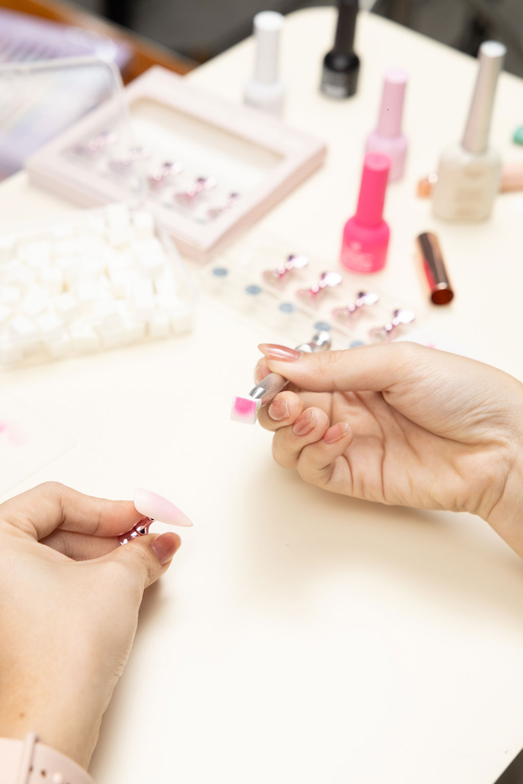 A person creating a pink ombre effect on a press-on nail using a small sponge applicator, with nail art supplies in the background