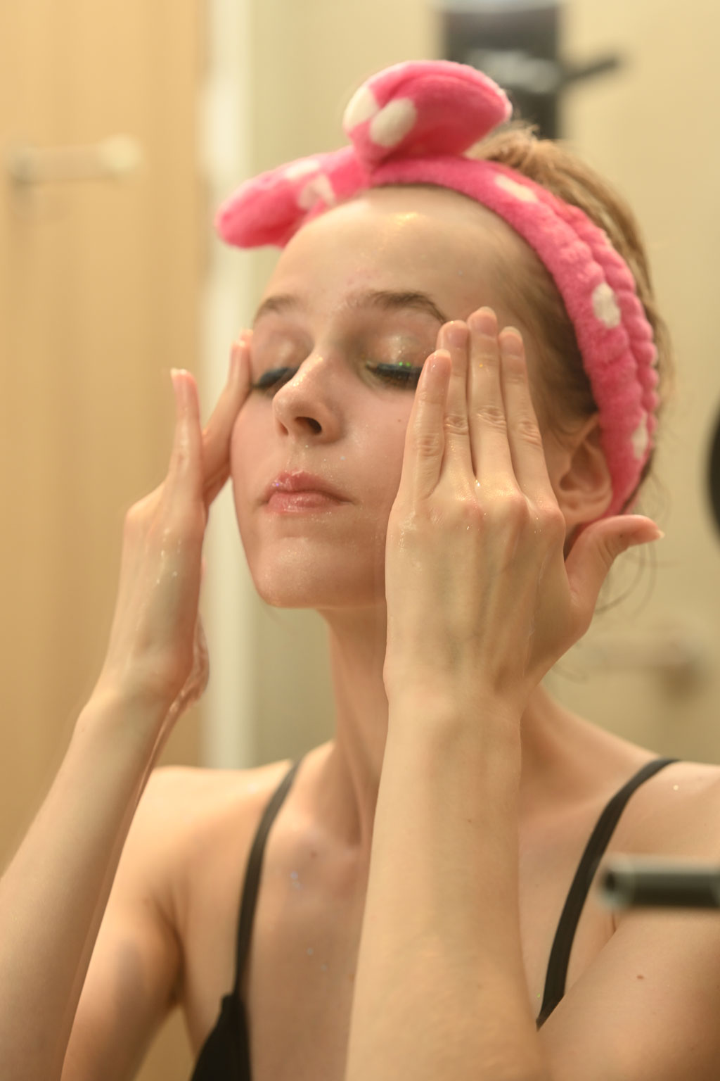 A young woman in a pink polka dot headband applying skincare.