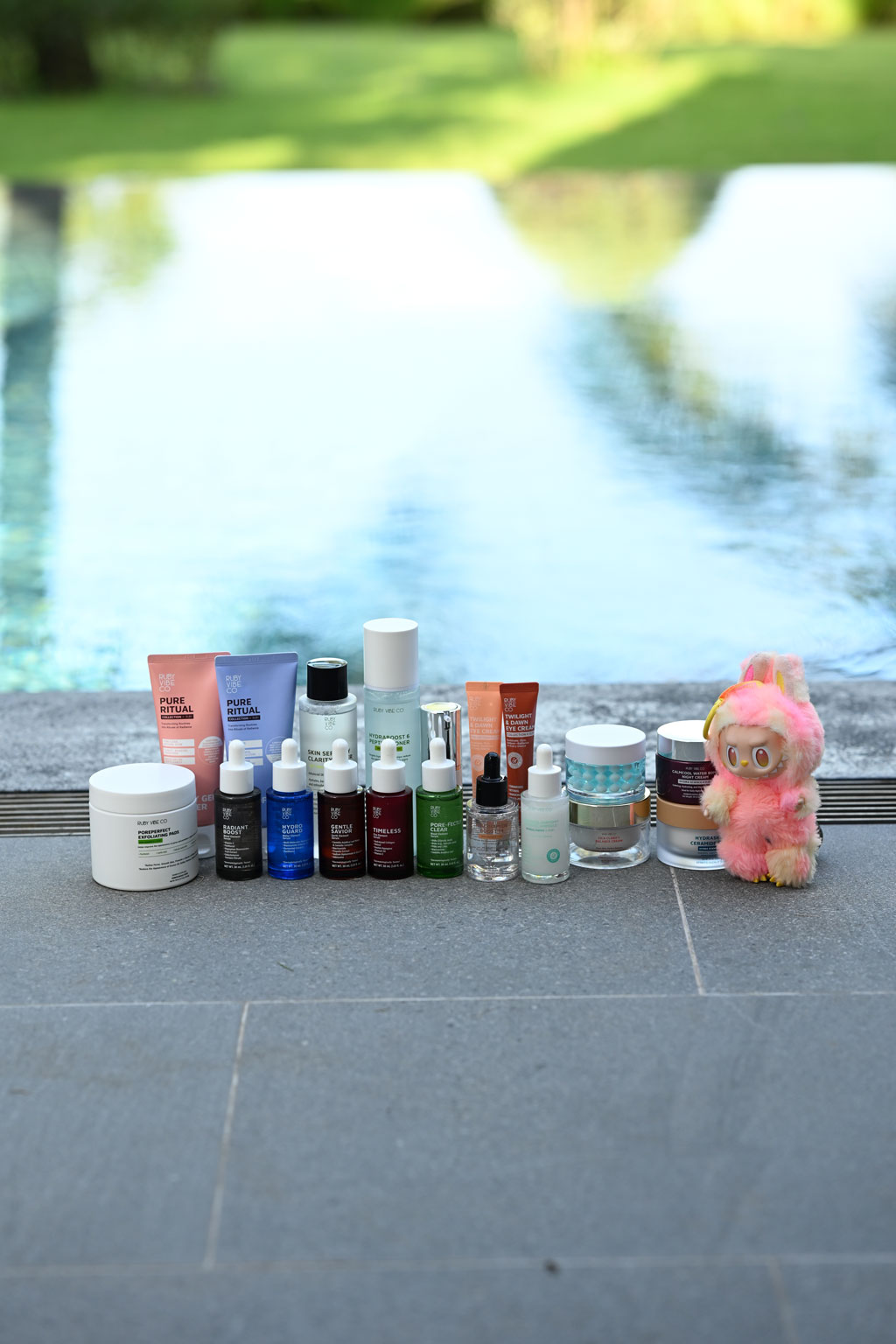 A product shot showcasing a full collection of skincare products. The items are arranged in a line on a grey poolside ledge, with the blue water of the pool creating a fresh, blurry background. A small, fluffy pink figurine sits at the end of the line.
