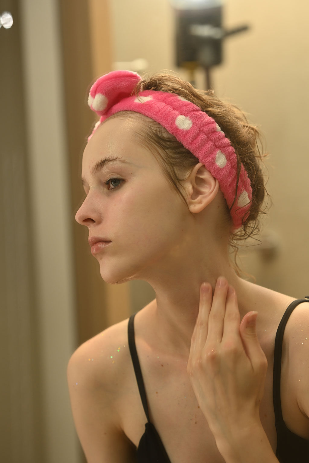 A young woman in a pink polka dot headband applying skincare.