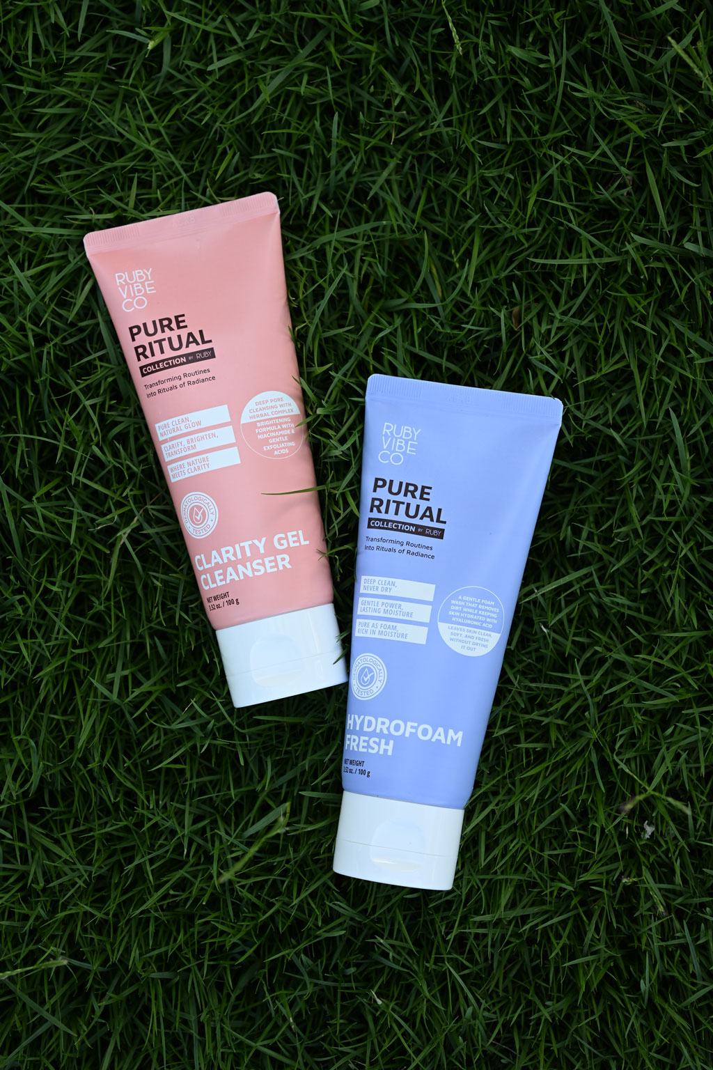 Two Ruby Vibe Co facial cleansers, the pink Clarity Gel and the blue Hydrofoam Fresh, lying on green grass.