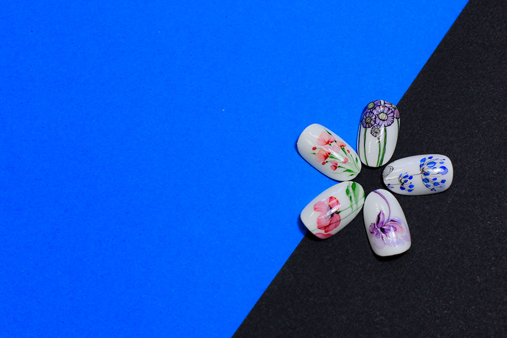 Five white artificial nail tips displaying various hand-painted floral designs, arranged in a flower shape on a blue and black background.