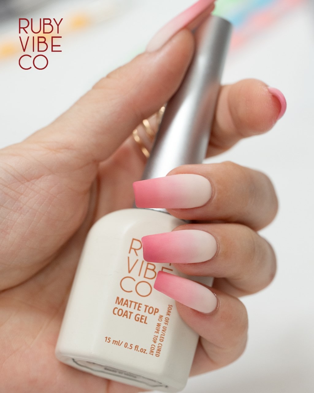 A hand with matte pink and white ombre nails holding a bottle of Ruby Vibe Co matte top coat gel.