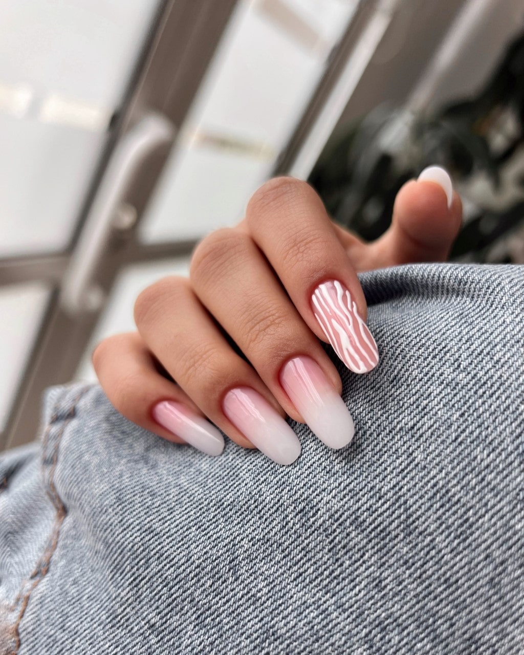 A hand with long, almond-shaped ombre nails, transitioning from pink to white, with a pink and white zebra-print accent nail, resting on blue denim.