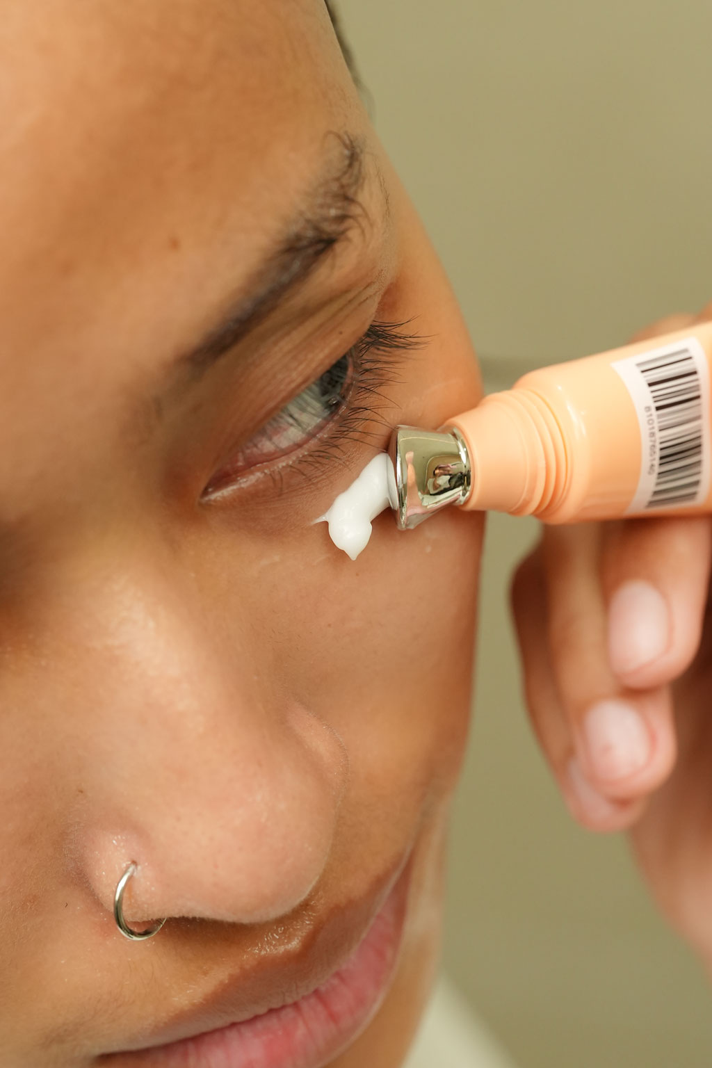  A close-up of a person applying white eye cream directly to their under-eye area from a peach-colored tube with a metal applicator tip.