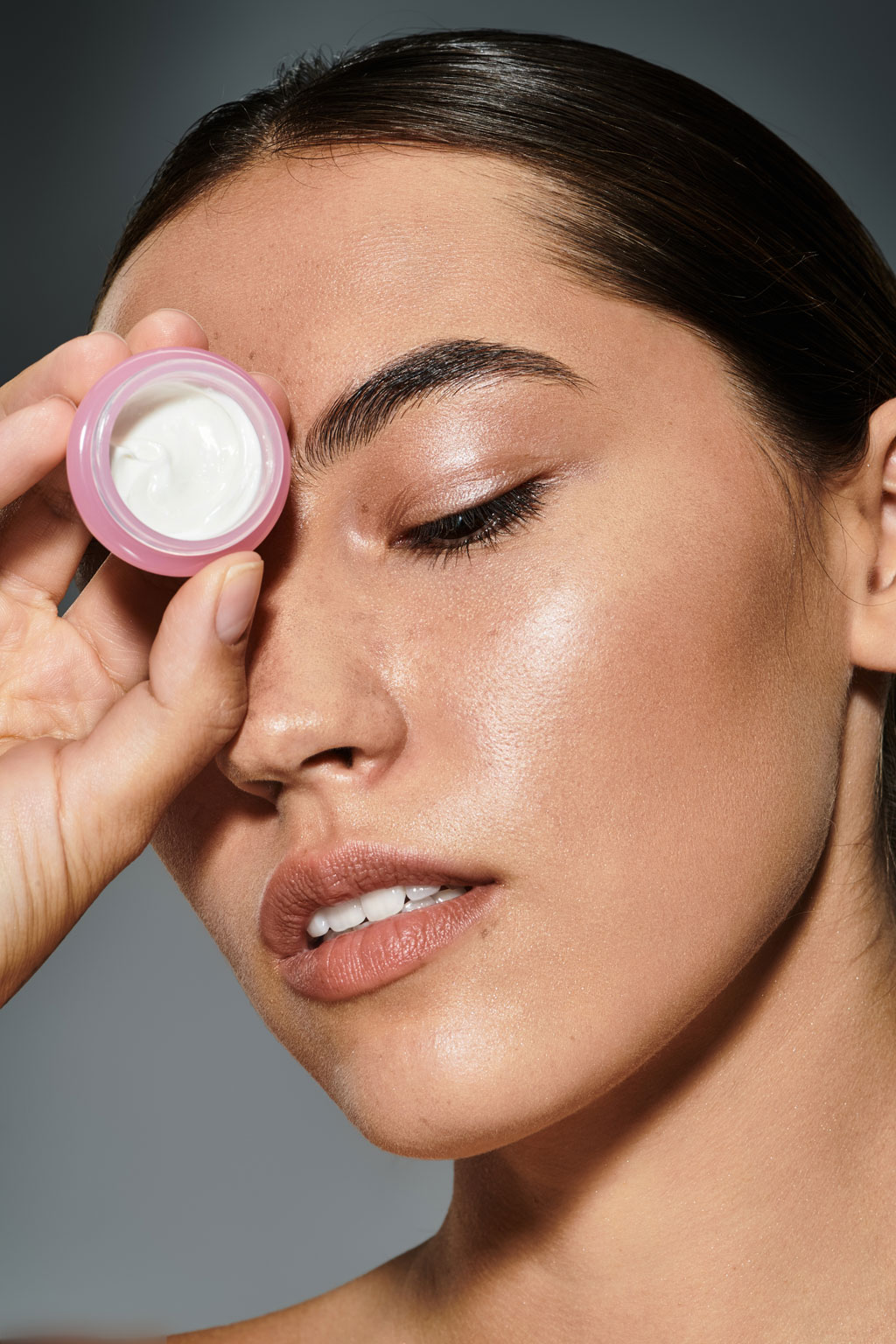 A beauty shot of a woman with glowing skin holding a small, open pot of white face cream over one eye, with her other eye closed.