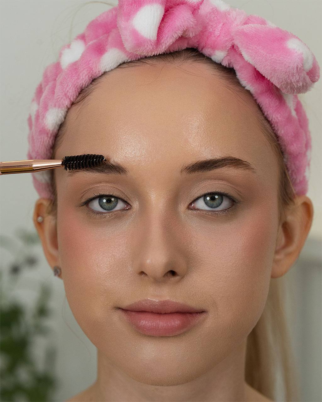 A close-up of a woman with blue eyes wearing a pink spa headband with white hearts while getting her eyebrows brushed with a spoolie.