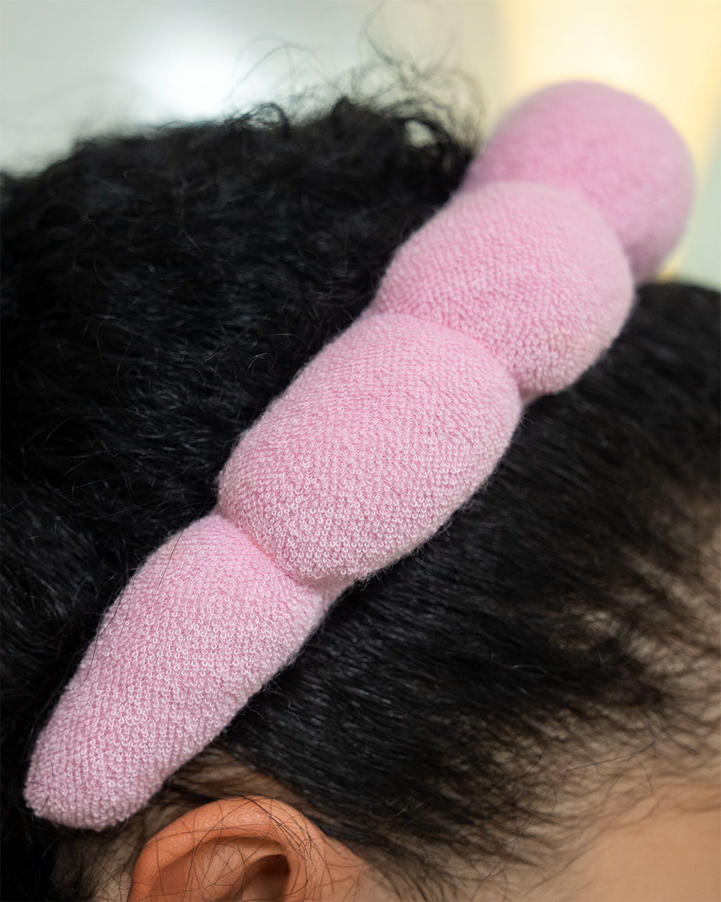 A close-up of a pink, puffy terry cloth spa headband being worn on dark, curly hair.