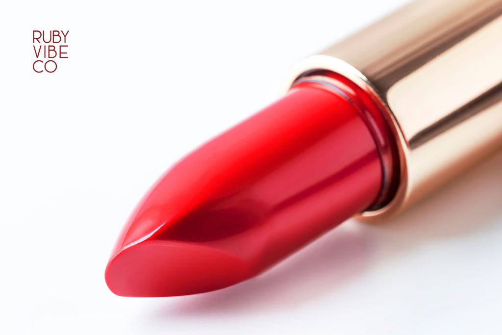 A macro photo of a vibrant, creamy red lipstick bullet emerging from its case against a clean white background.