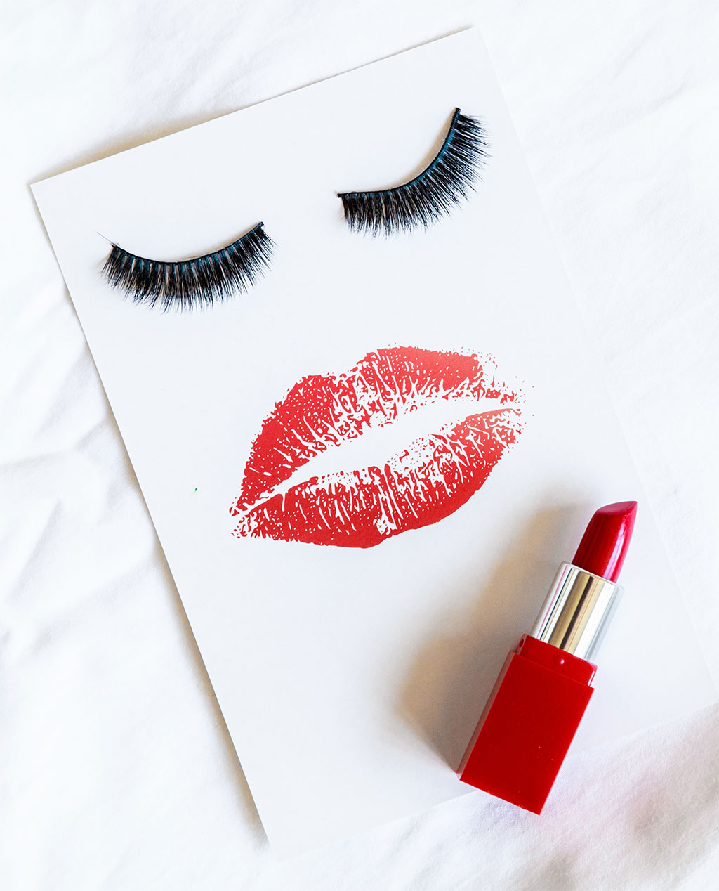 A creative flat lay on a white sheet featuring a red lipstick tube next to a card with a face made of false eyelashes and a red lipstick kiss print.