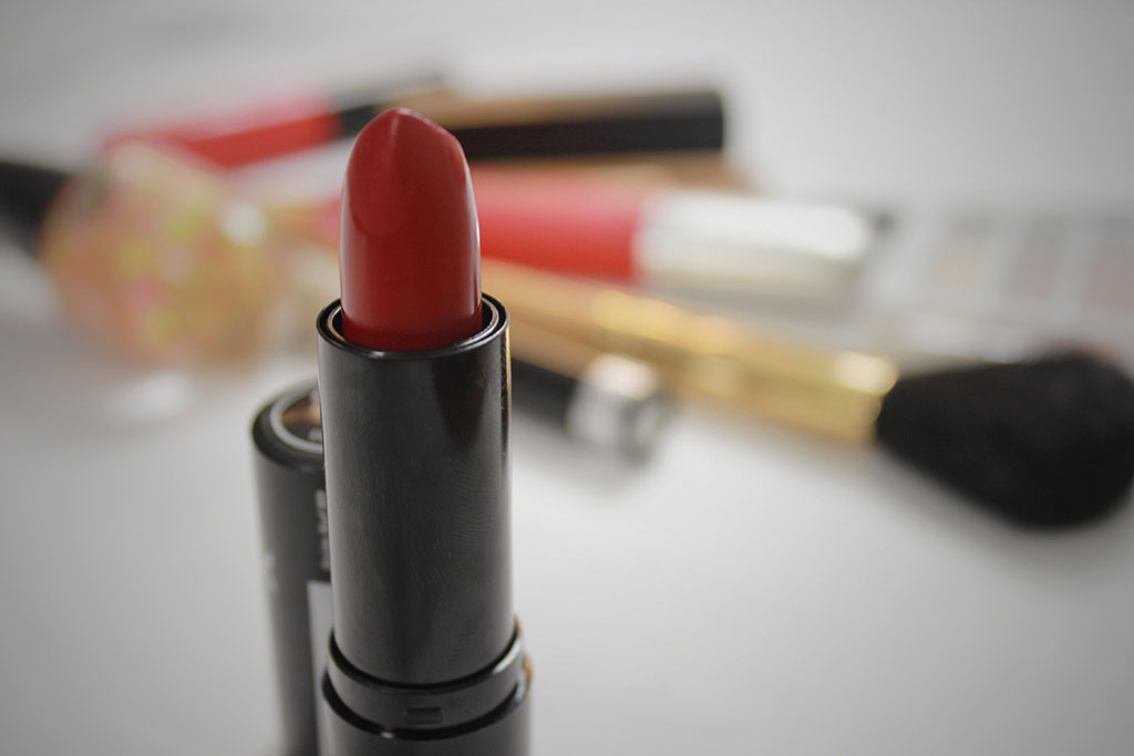 An open tube of red lipstick in a black case, positioned in the foreground with various other makeup items like brushes and lip gloss blurred in the background.