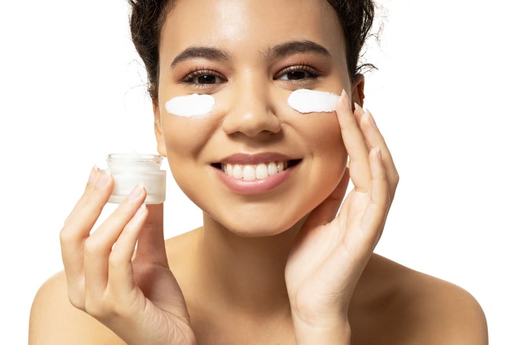 A smiling woman looking at the camera while holding a jar of cream and applying dabs of it under her eyes with her finger. 