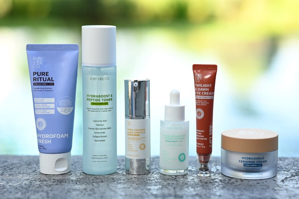 A lineup of various skincare products, including a cleanser, toner, serum, and creams, displayed on a ledge with a blurred natural background.